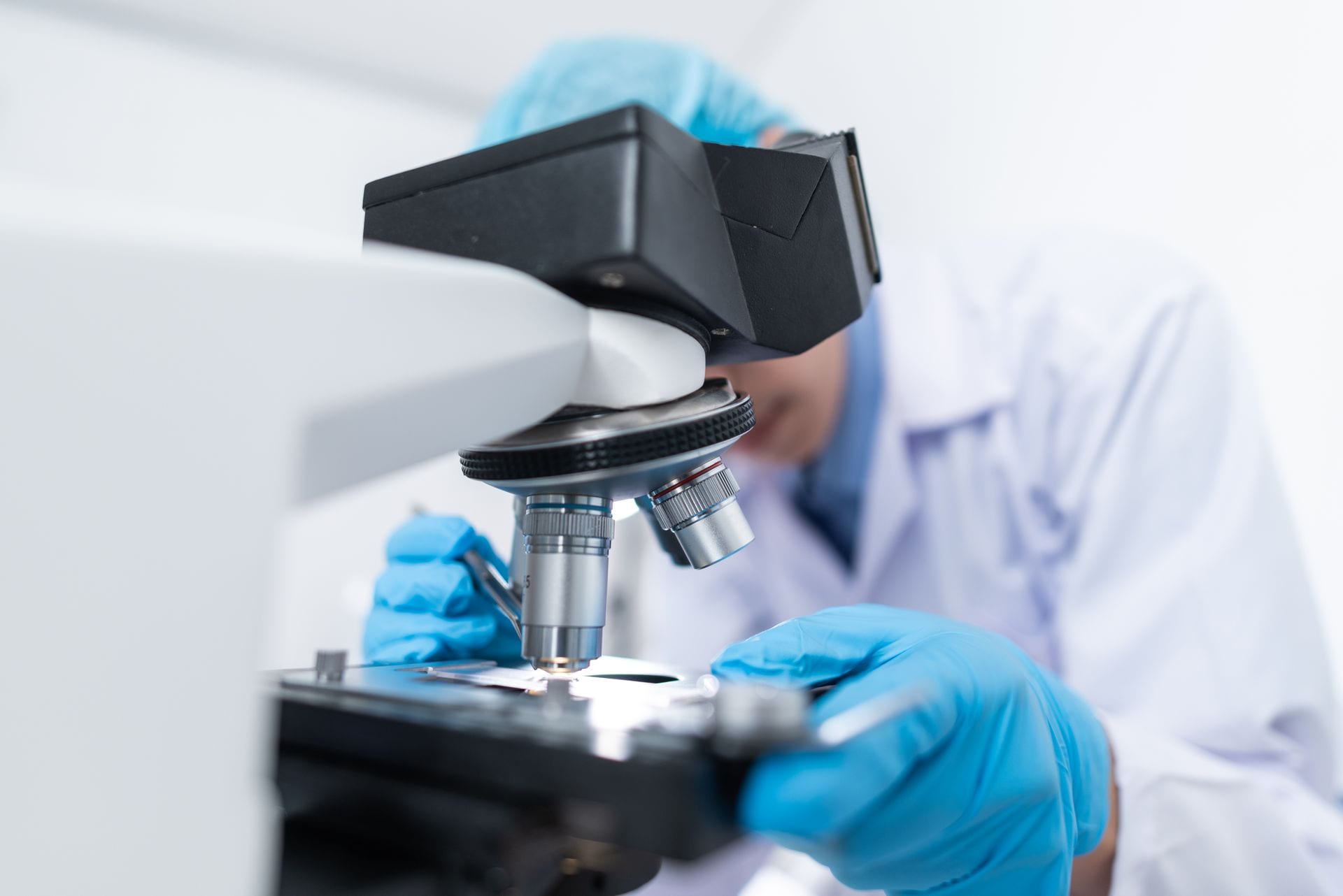 Una persona con bata blanca y guantes azules observa una muestra en un microscopio, en primer plano el objetivo y la platina.