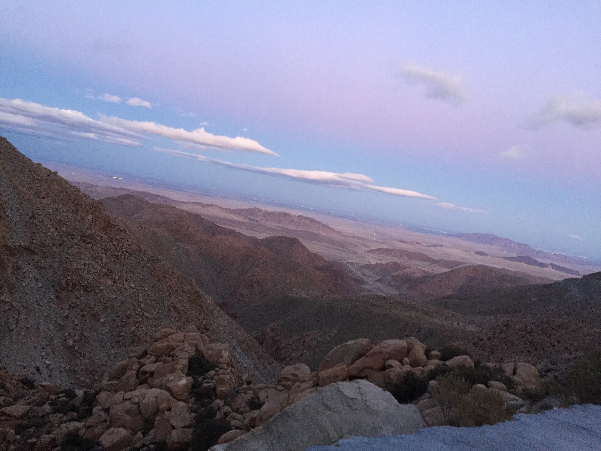 Vista al atardecer desde una sierra rocosa hacia un valle desértico, bajo un cielo con nubes alargadas.
