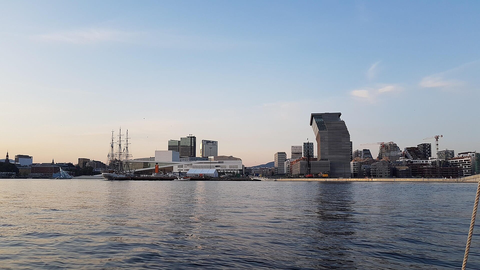 View of Oslo from the sea at sunset, with the Opera House, office buildings and a tall ship moored at the quay.