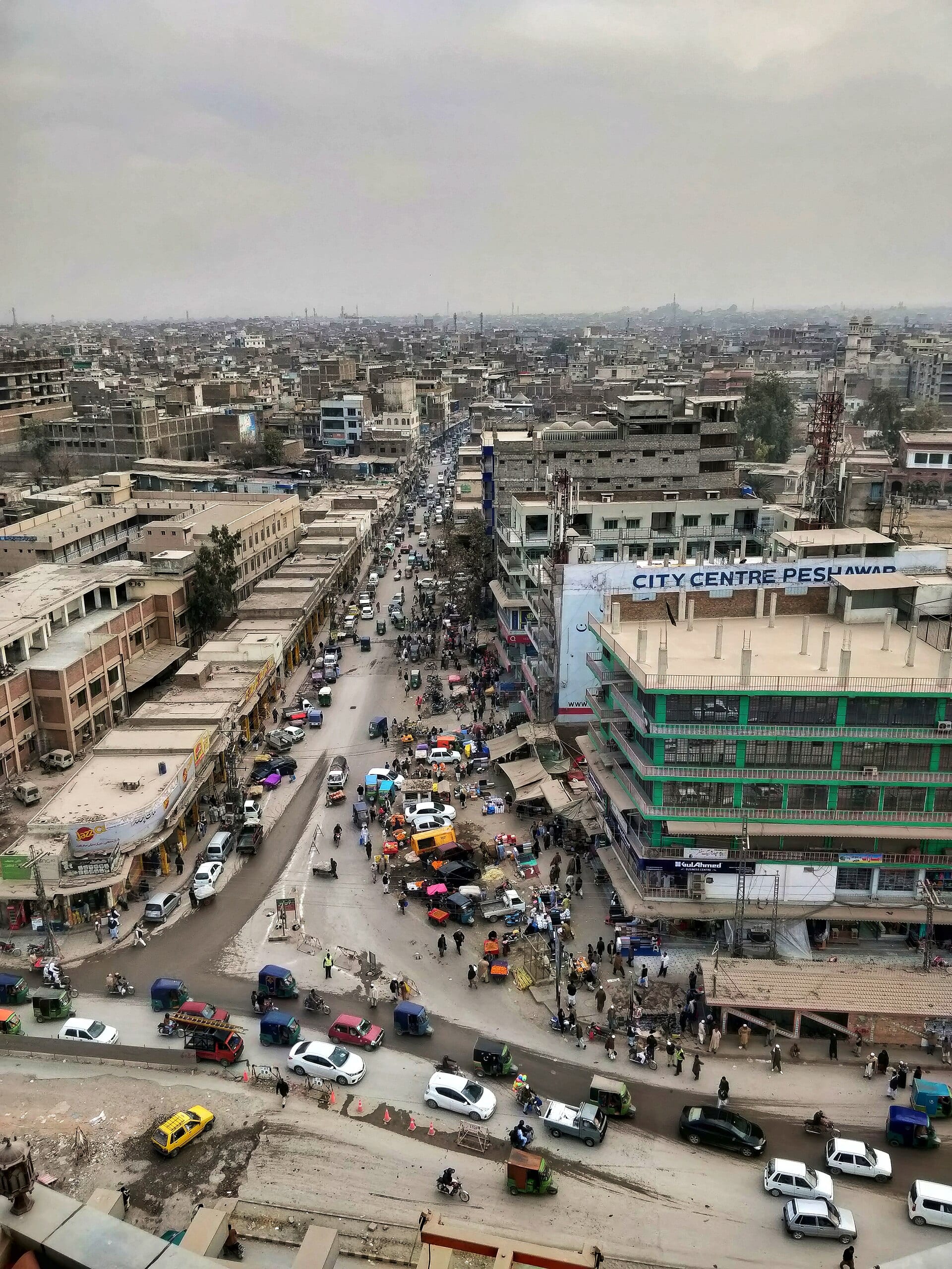 Aerial view of a busy avenue in Peshawar, with auto rickshaws, shops and a green multistory building.