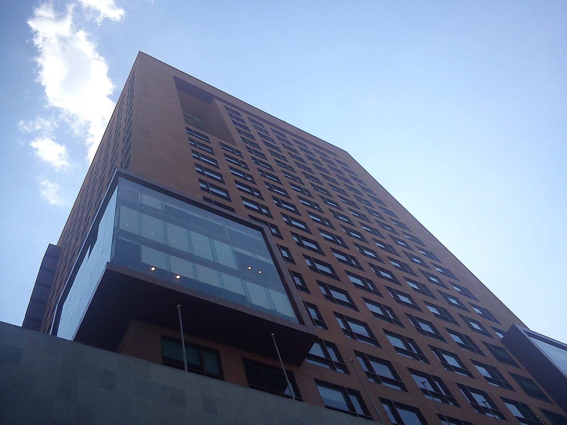 Torre de ladrillo rojizo con ventanas en hileras y un voladizo de cristal, vista desde abajo contra un cielo azul con una nube.