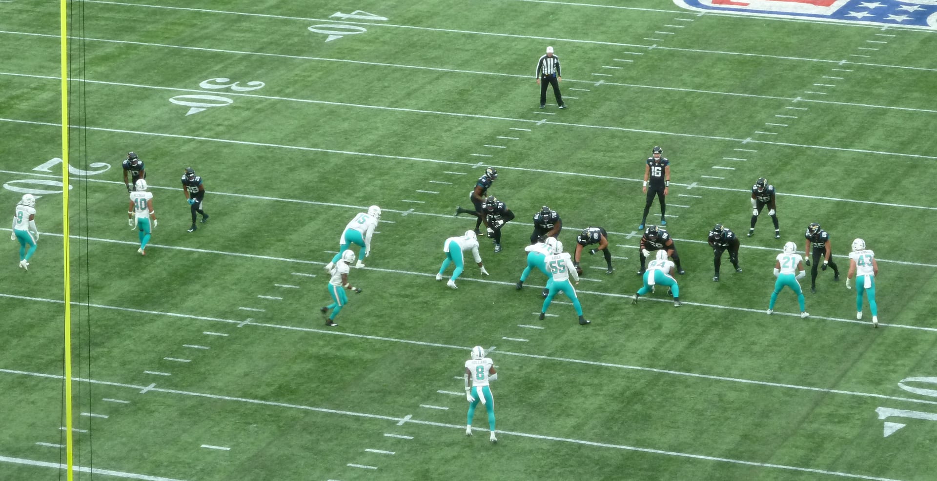 Jugadores de Jaguars con uniforme oscuro y de Dolphins con uniforme blanco y turquesa alineados antes de una jugada sobre el césped del estadio, con un árbitro al fondo.