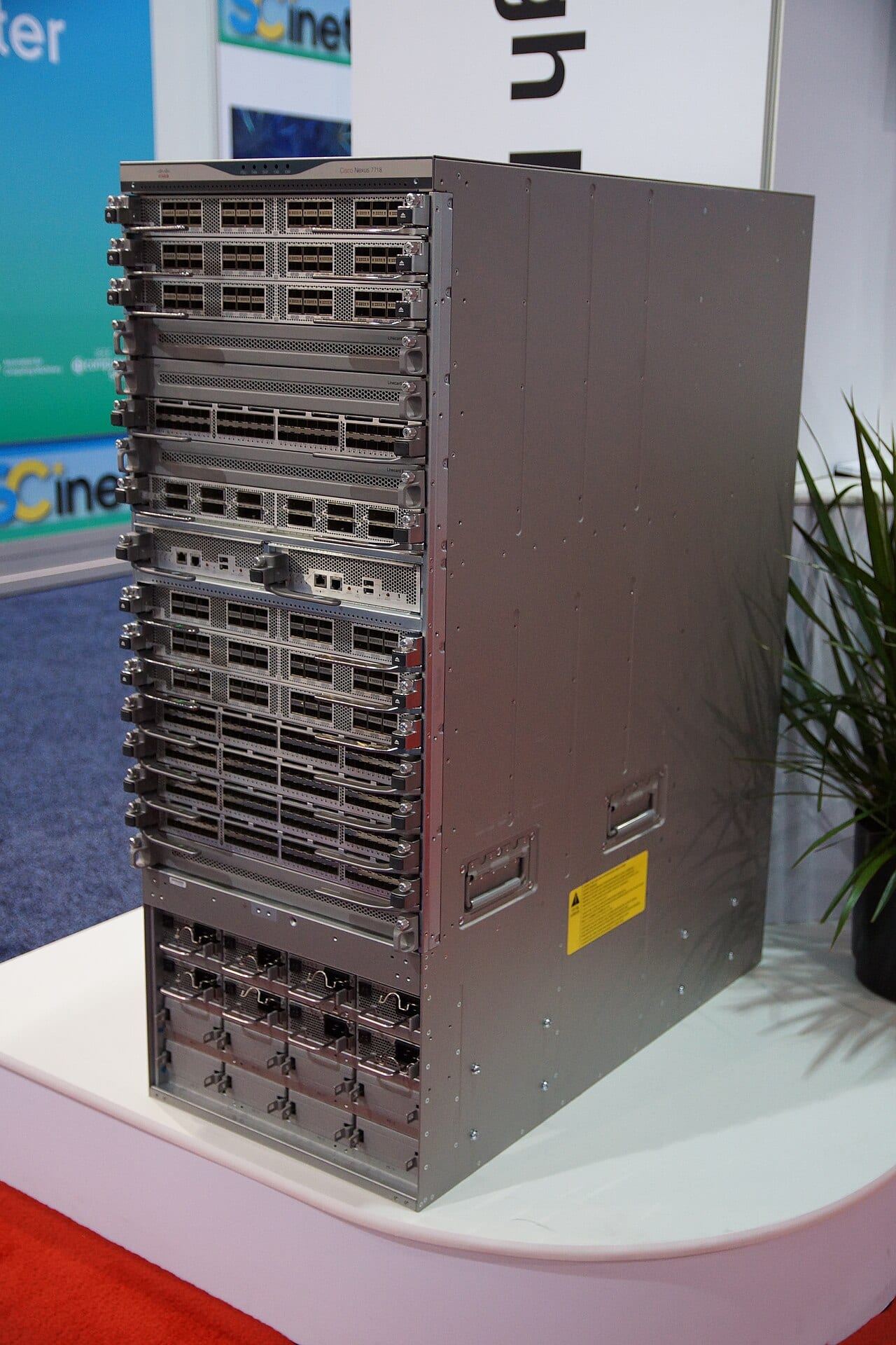 Gray chassis of a Cisco Nexus 7718 switch with stacked port modules, displayed on a white platform next to a plant.