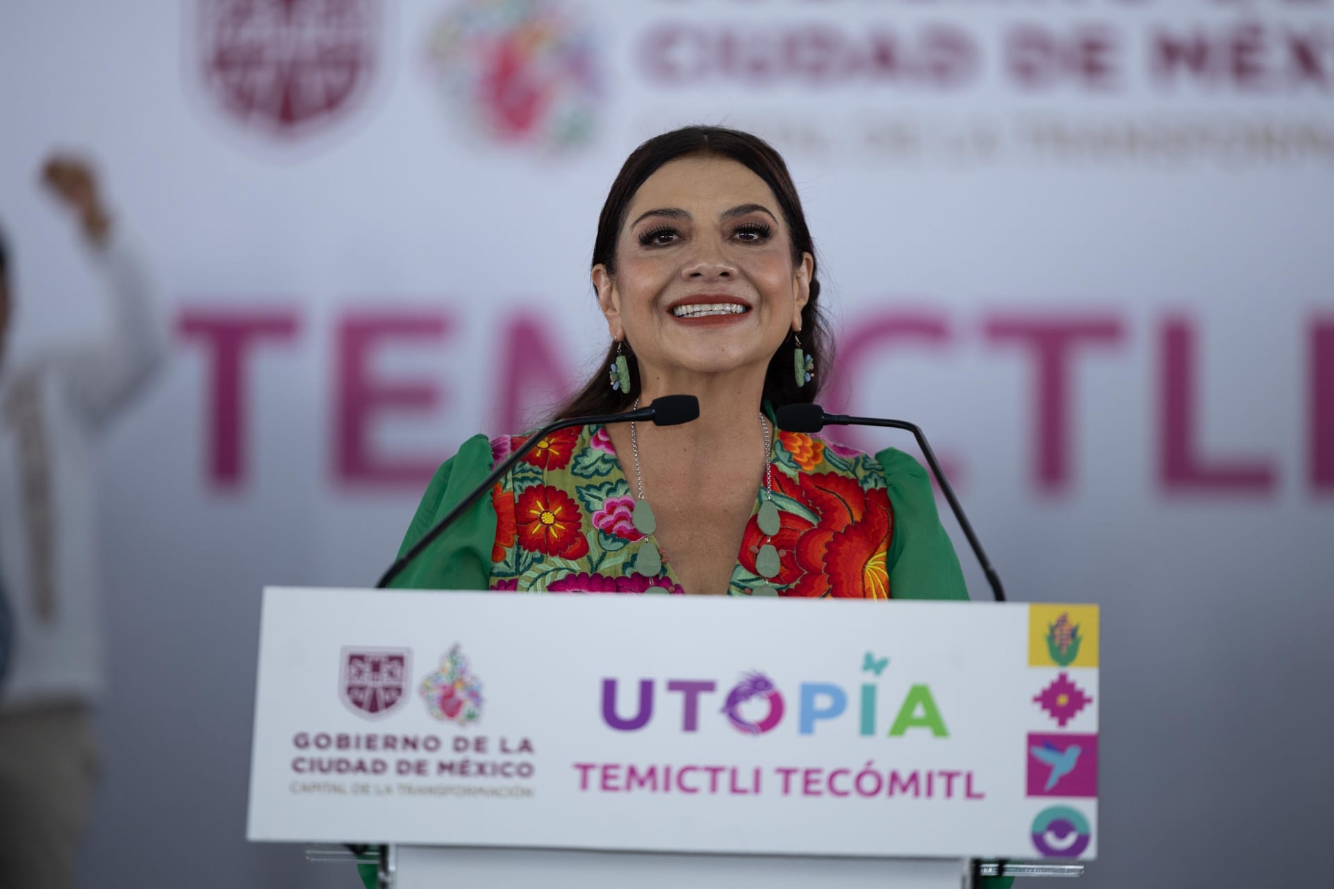 Clara Brugada sonríe frente a dos micrófonos en un podio con el logotipo de Utopía Temictli Tecómitl y un fondo con el nombre del recinto.