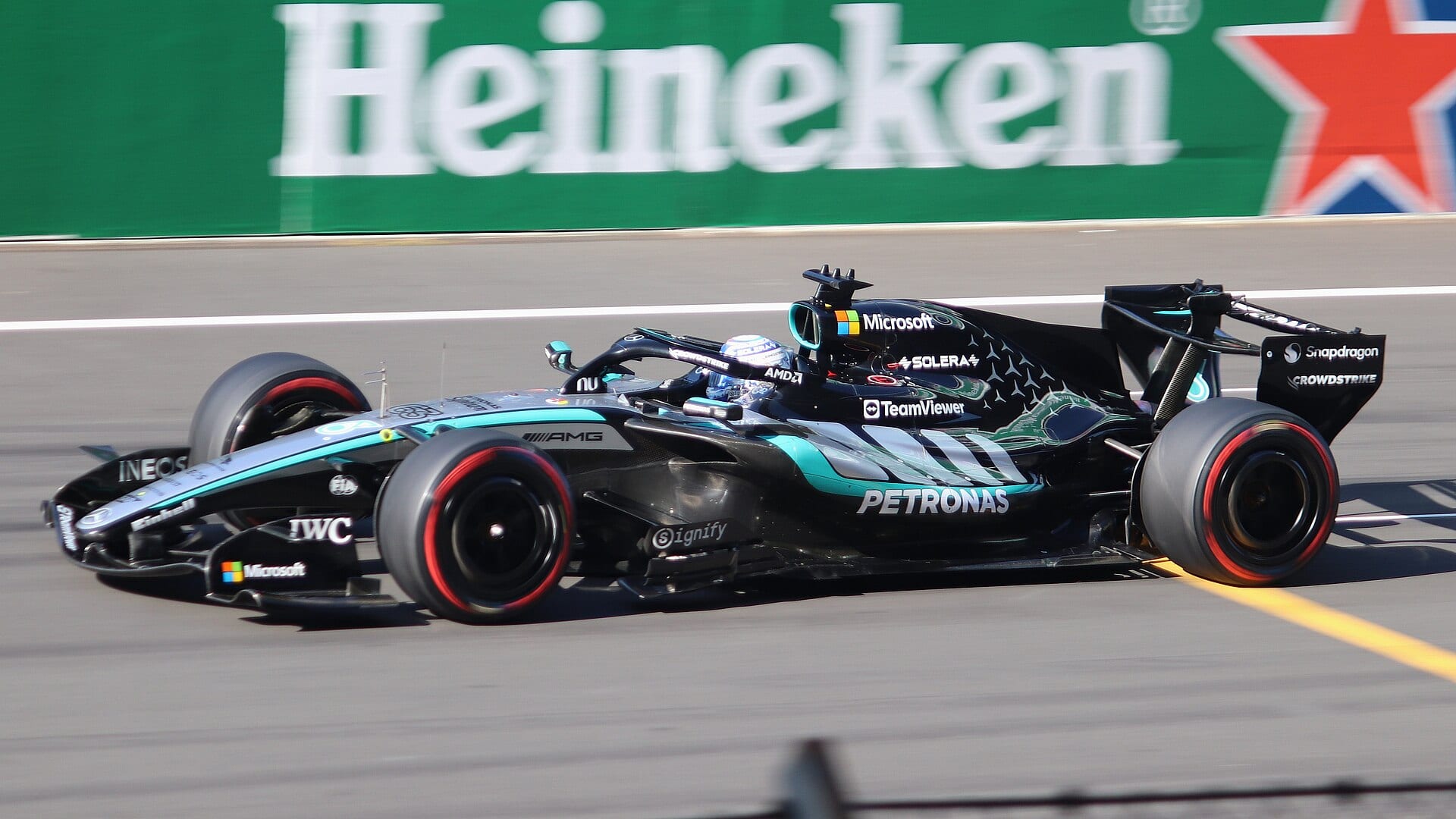 Un monoplaza negro con detalles turquesa de Mercedes pasa a toda velocidad frente a un muro verde de publicidad.