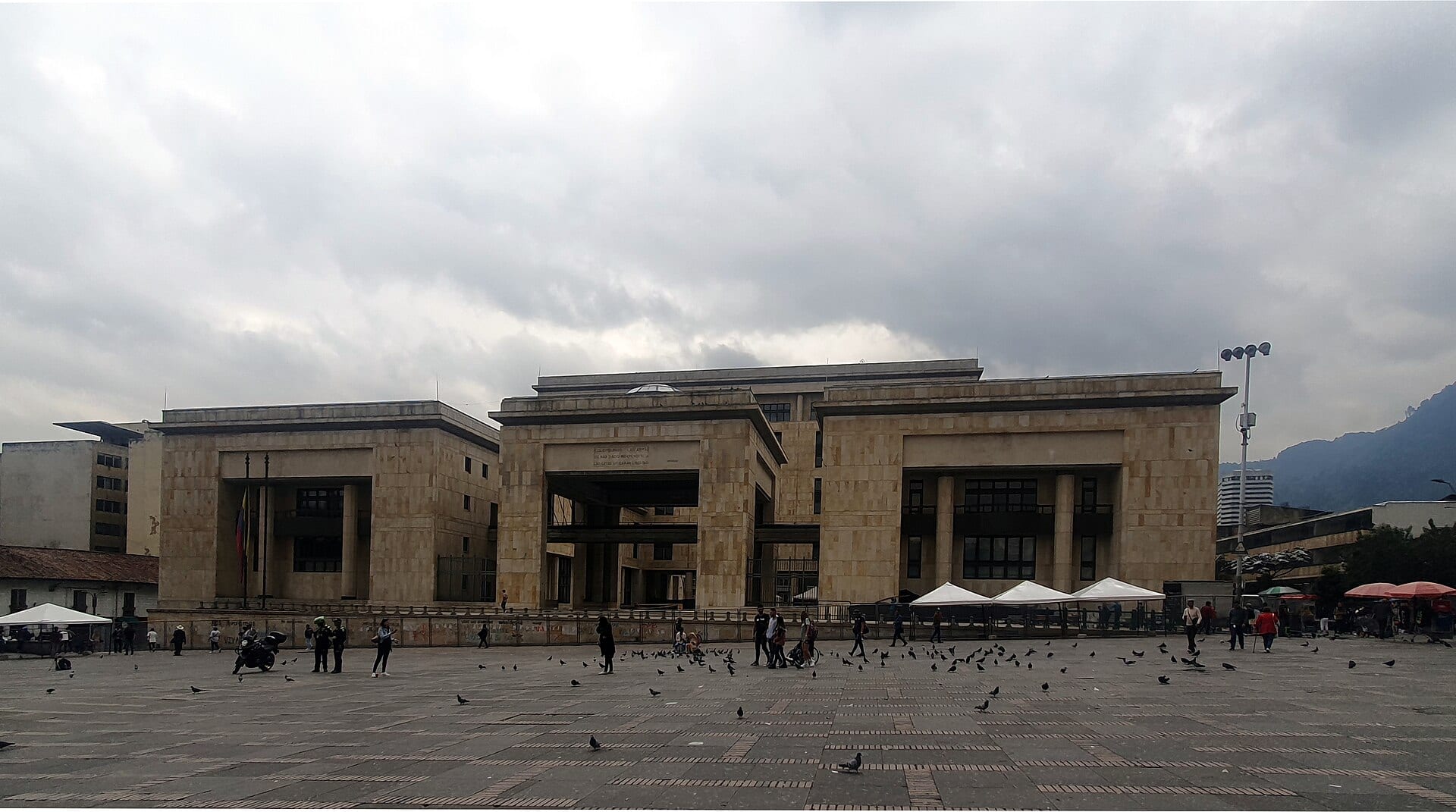 Fachada de piedra clara del Palacio de Justicia en la Plaza de Bolívar de Bogotá, con palomas y transeúntes en primer plano.