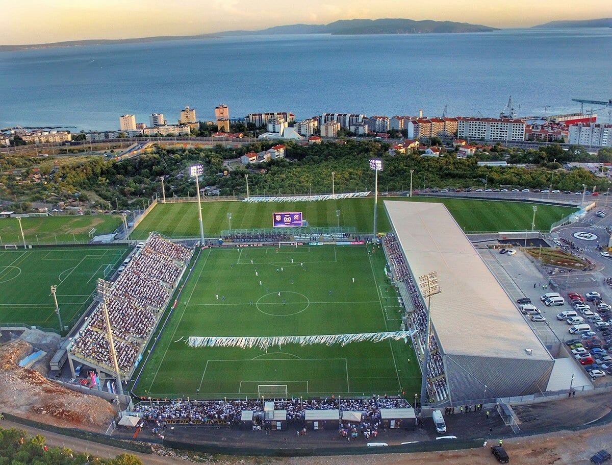 Vista aérea del estadio Rujevica de Rijeka durante un partido, con las tribunas ocupadas, el campo iluminado y el mar Adriático al fondo.
