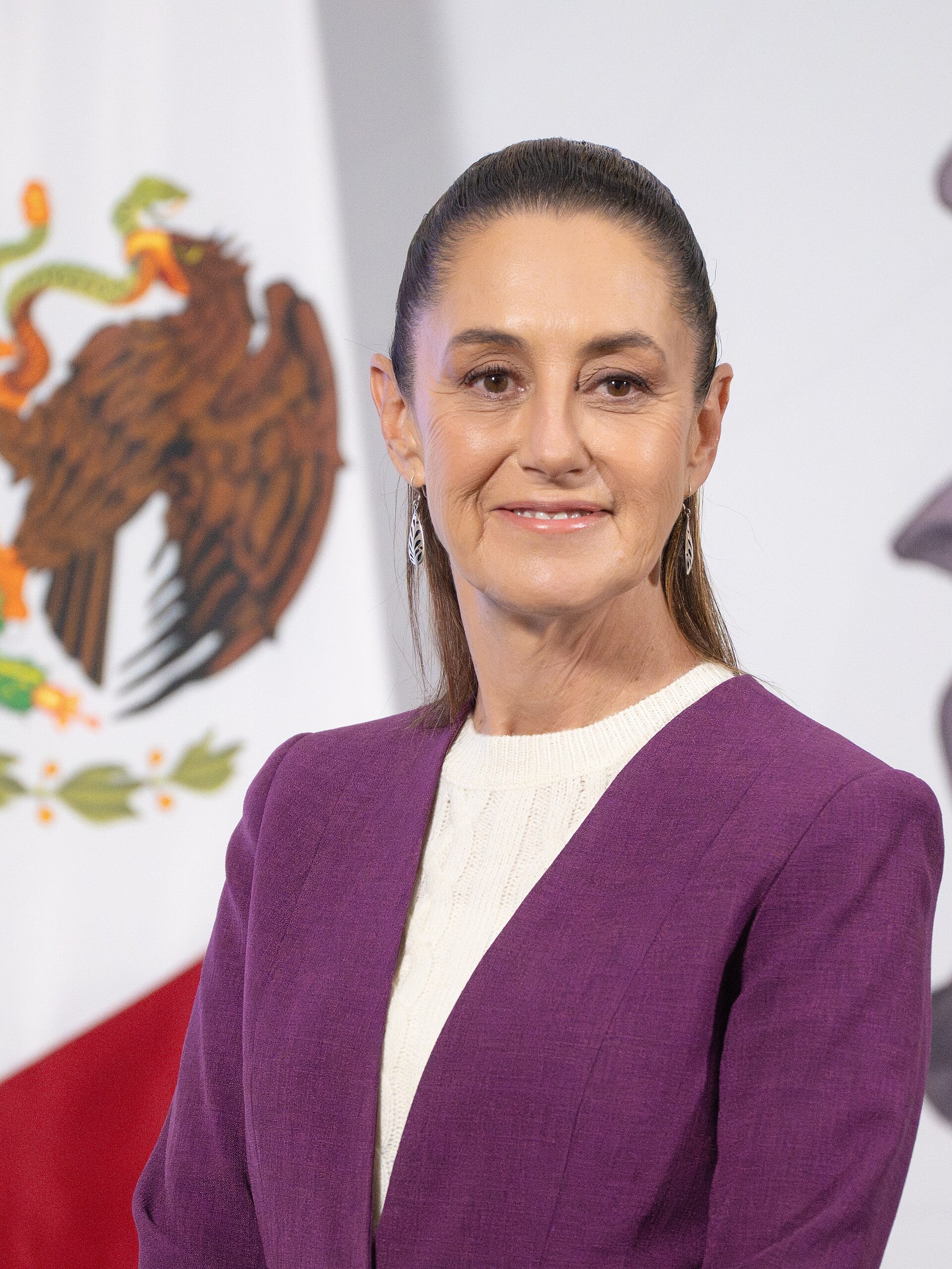 Retrato de Claudia Sheinbaum con saco morado y suéter blanco, frente a una bandera de México con el escudo nacional.