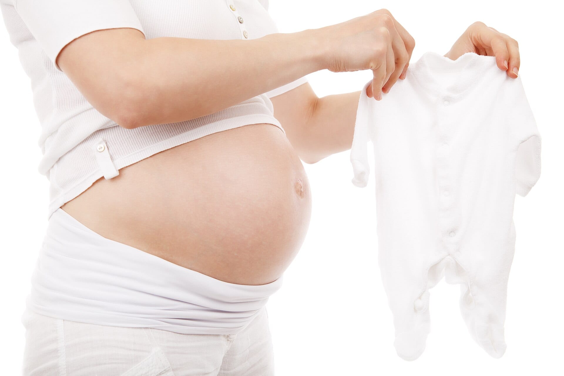 Torso de una mujer embarazada, vestida de blanco, que sostiene con las dos manos una prenda de bebé también blanca.