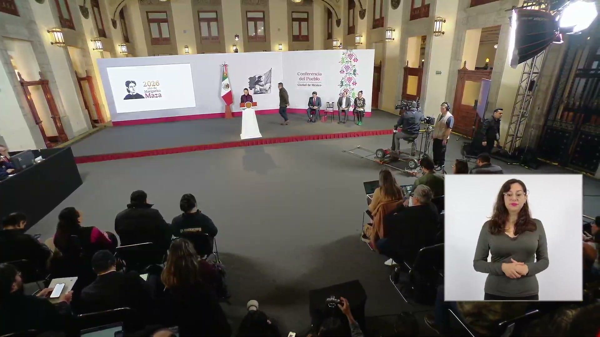 Vista general del salón de la conferencia matutina del 19 de octubre de 2026, con la presentadora en el podio y una intérprete de lengua de señas.