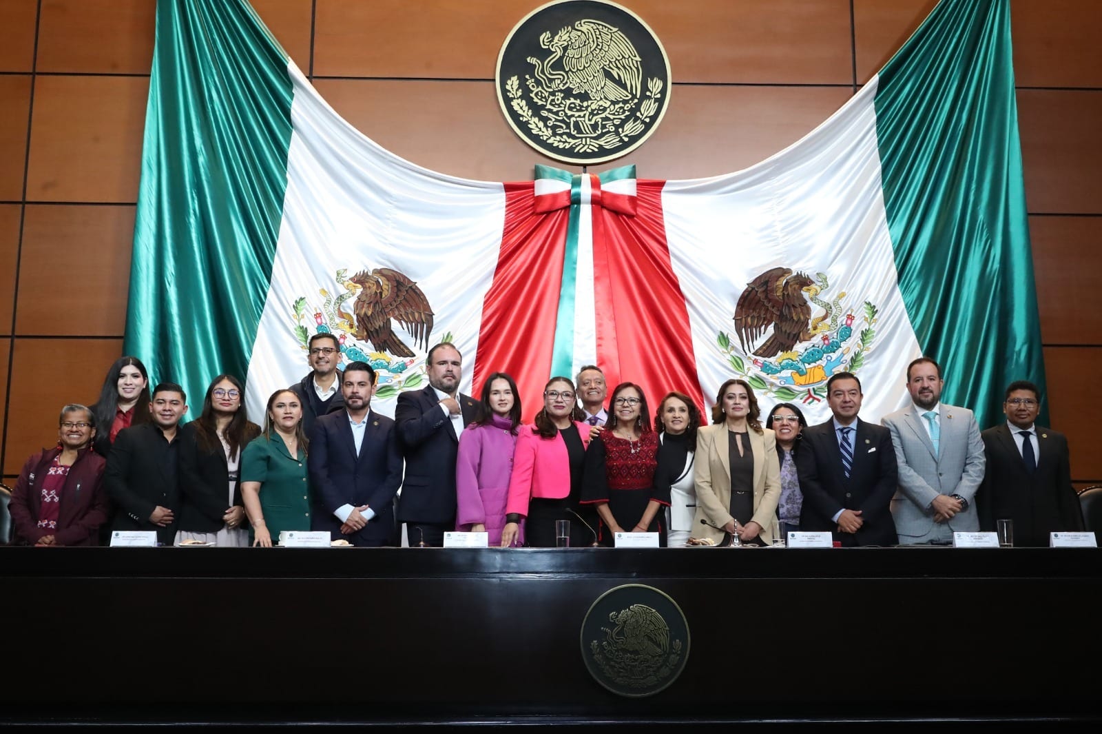 Cerca de veinte legisladores y la secretaria de Bienestar posan de pie detrás de la mesa de sesiones, bajo la bandera y el escudo nacional.