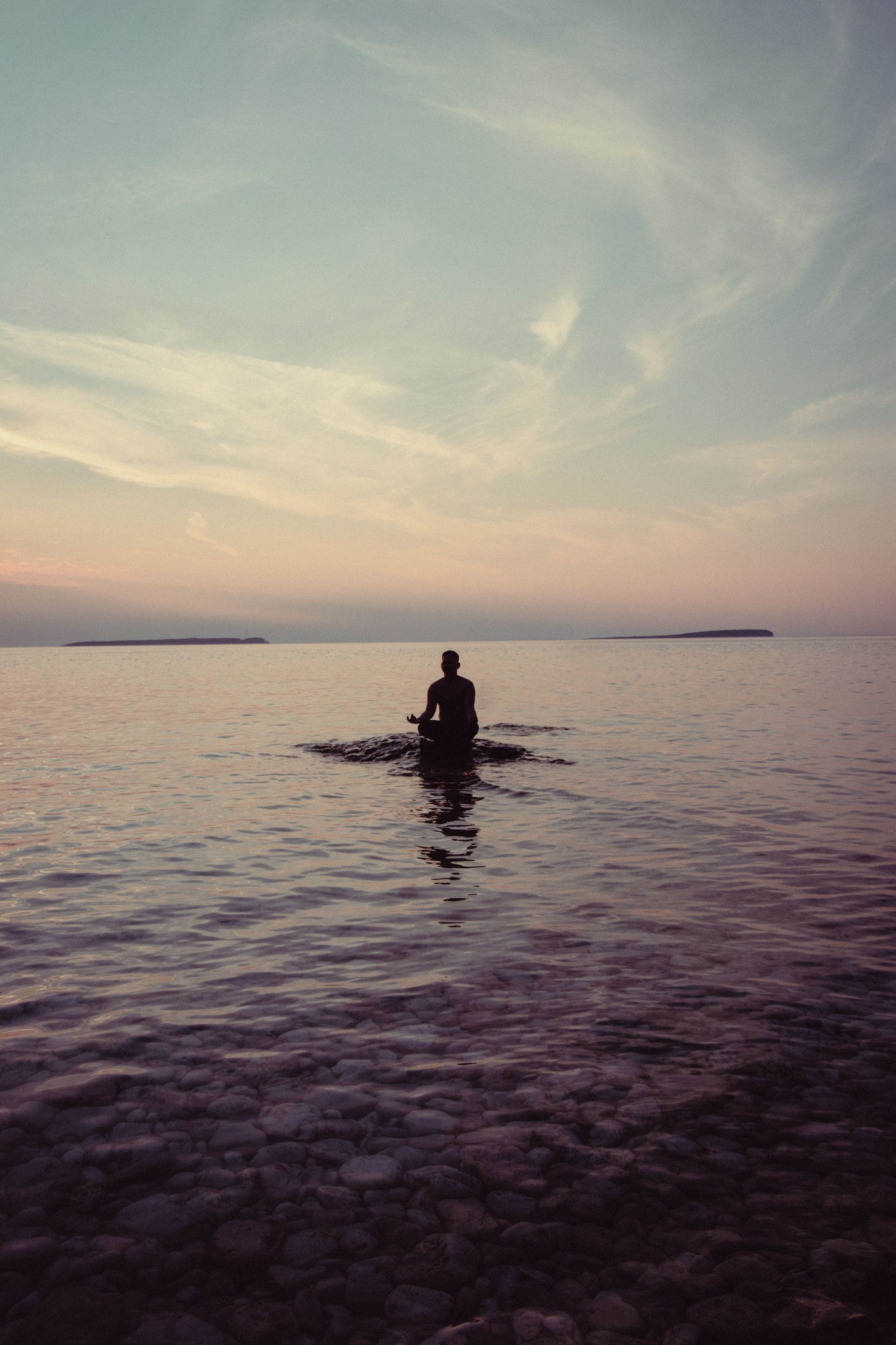 La silueta de una persona sentada en postura de meditación sobre una roca en el agua, con el mar y el cielo al atardecer.
