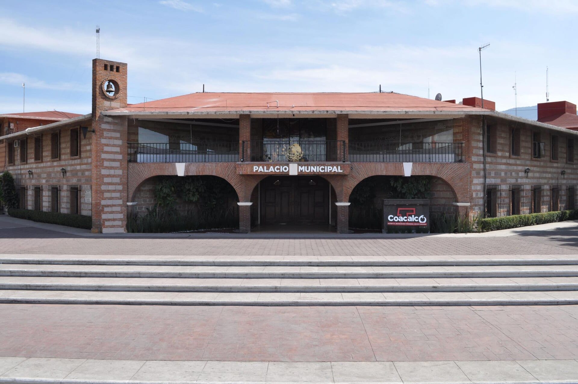 Fachada de ladrillo rojo del Palacio Municipal de Coacalco, con arcos, un balcón y una plaza de adoquín al frente.