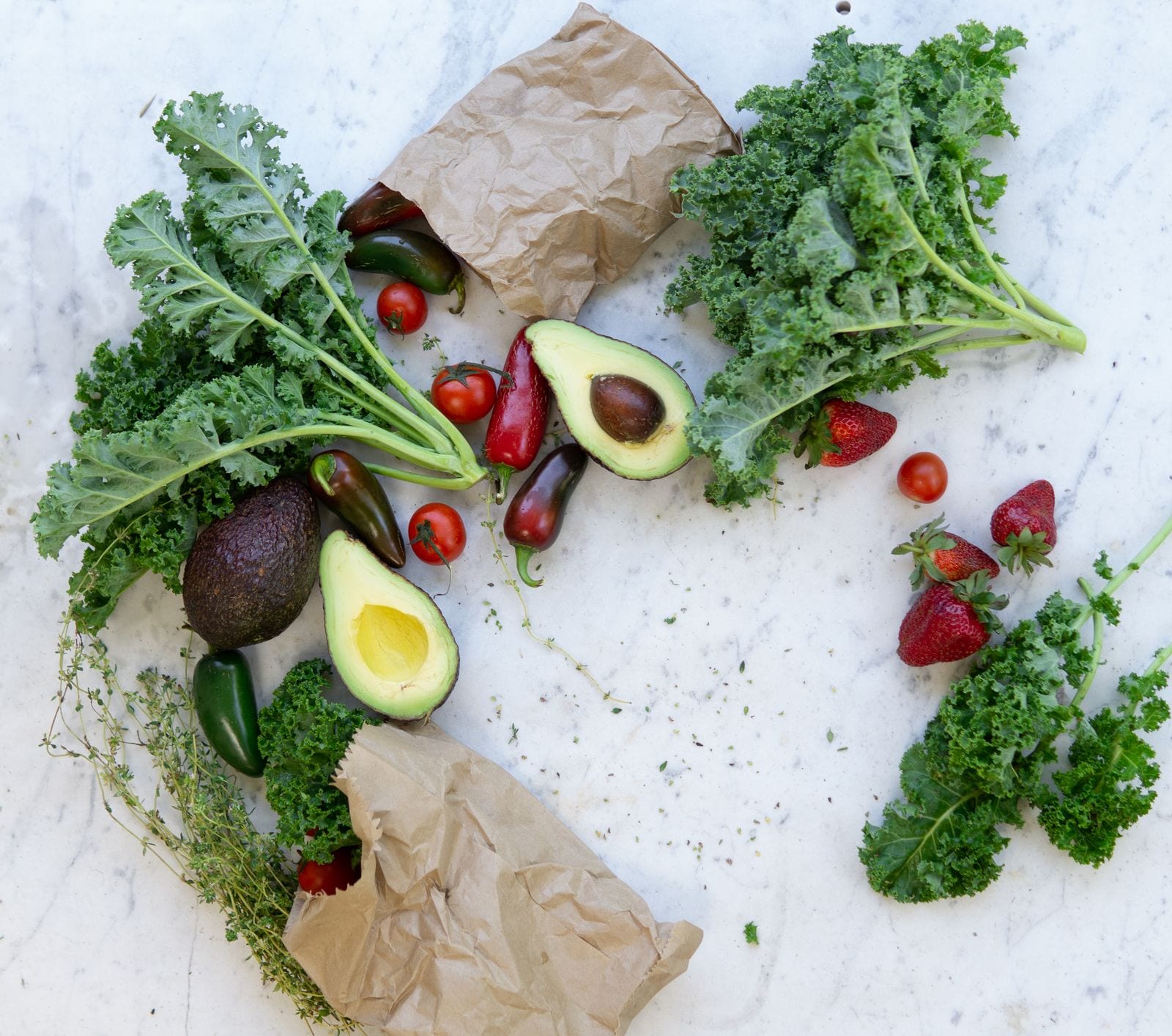 Col rizada, aguacates, jitomates, chiles y fresas sobre una mesa de mármol blanco, con dos bolsas de papel.