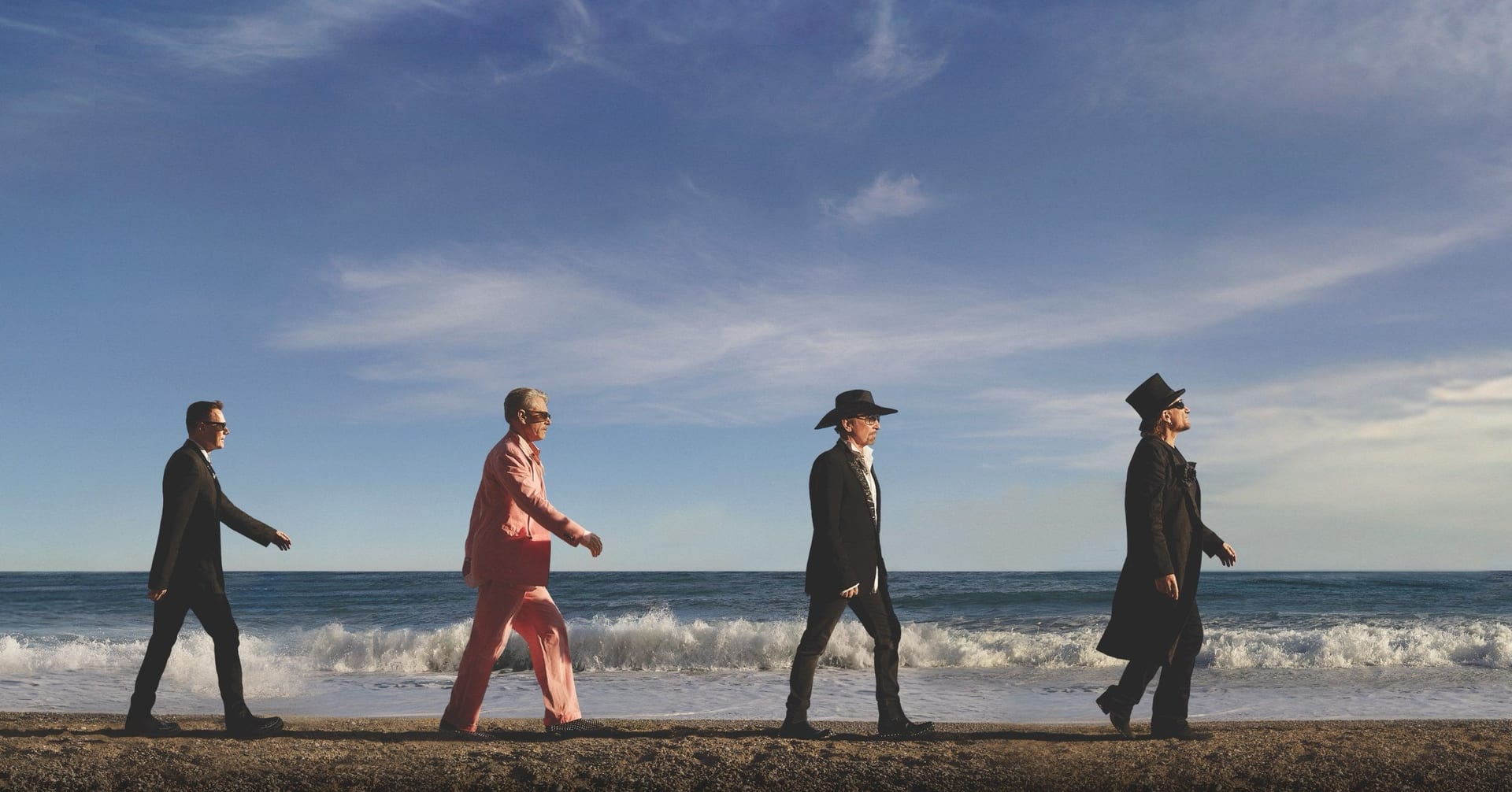 The four members of U2 walk in single file along a gravel beach with waves behind them, in dark suits, one in pink, some wearing hats.