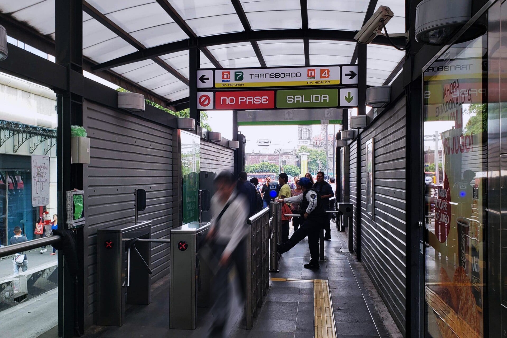 Acceso a la estación Hidalgo con torniquetes, un letrero de transbordo al Metro y una franja amarilla de guía táctil en el piso.