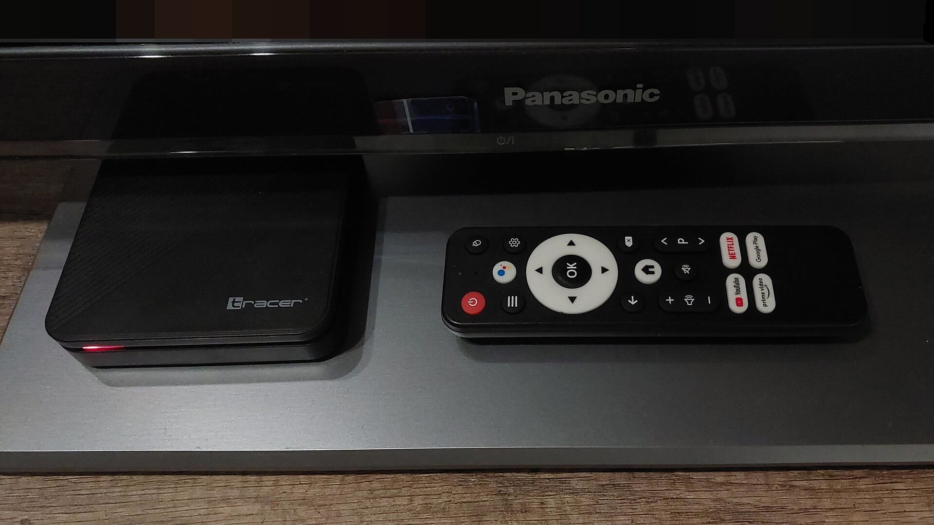 Una caja negra con sistema Android TV junto a su control remoto, sobre un mueble bajo un televisor.