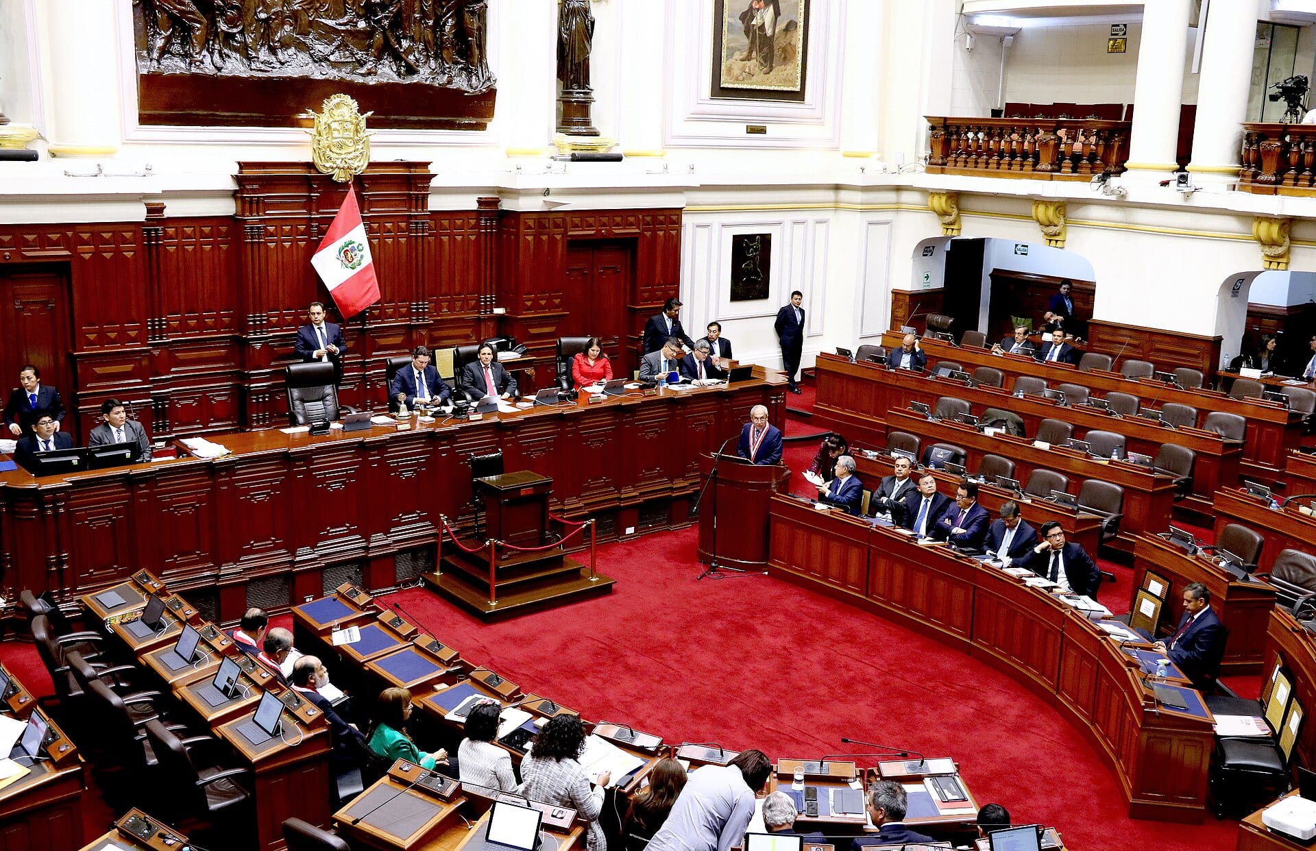 Una sesión en el hemiciclo del Congreso de Perú, con una bandera peruana junto a la mesa directiva y un orador en el podio.