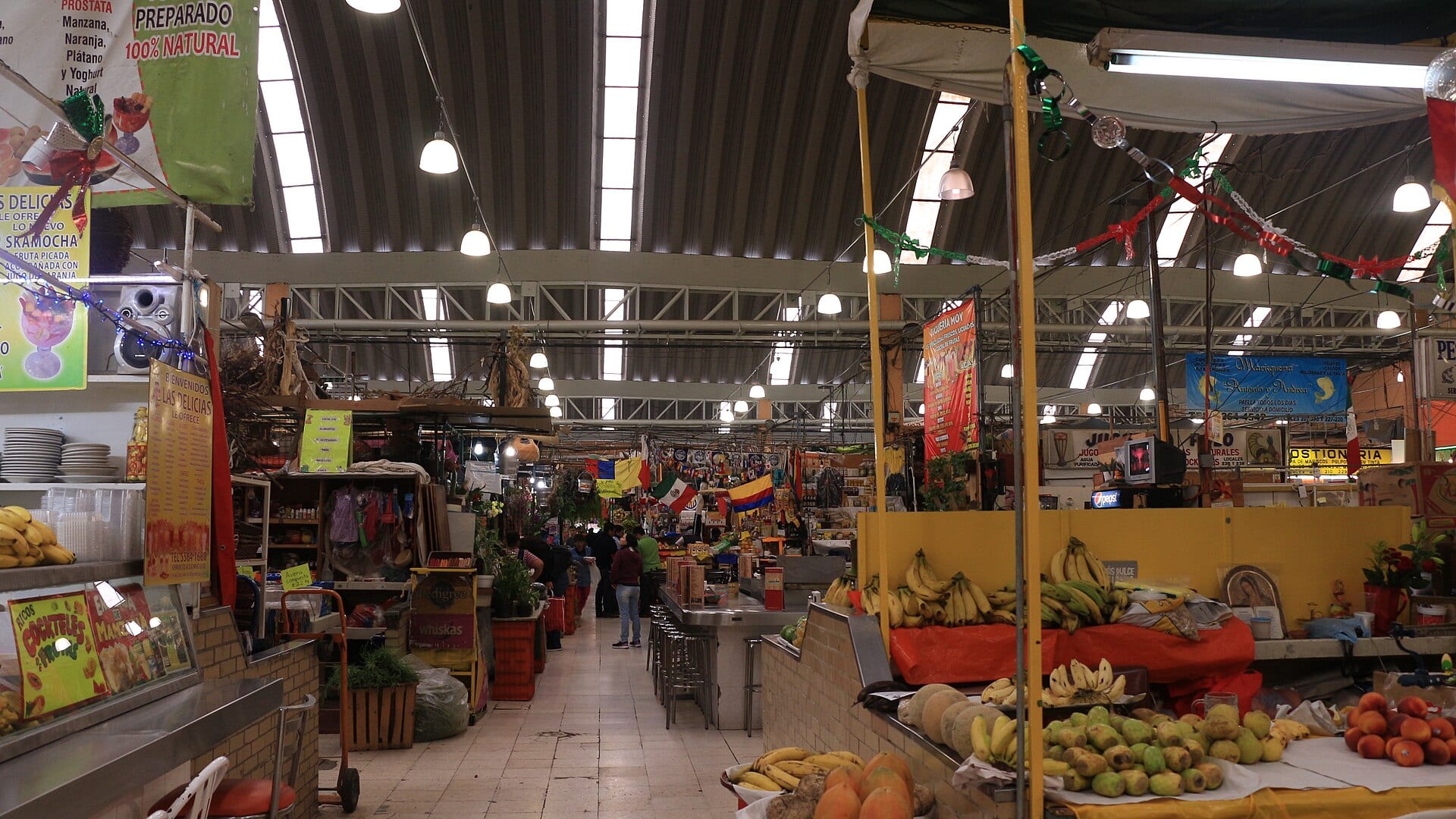 Pasillo central de un mercado techado con puestos de fruta a los lados y luminarias colgadas de una bóveda metálica.