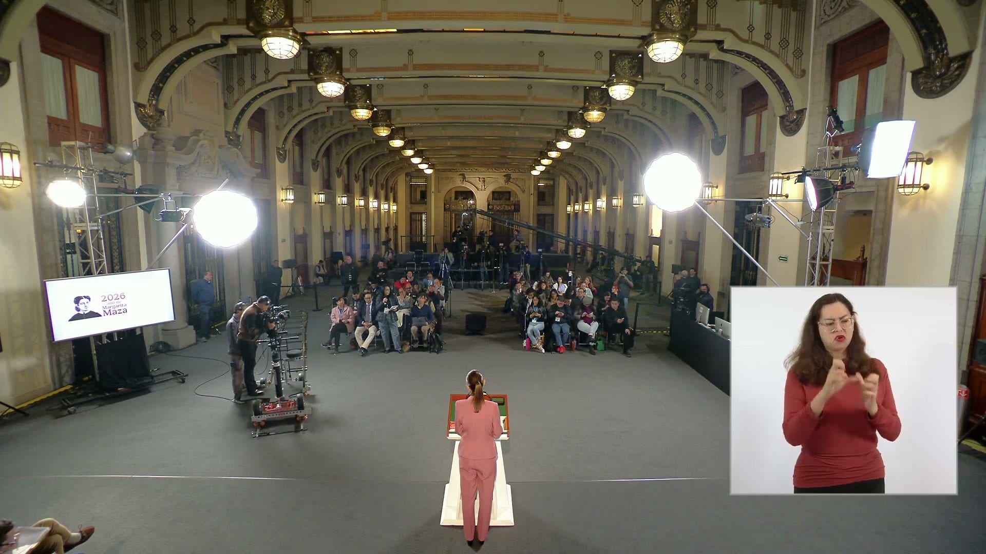 Vista desde atrás de la presidenta en el podio del salón de la conferencia, con reporteros sentados al fondo y una intérprete de lengua de señas a la derecha.