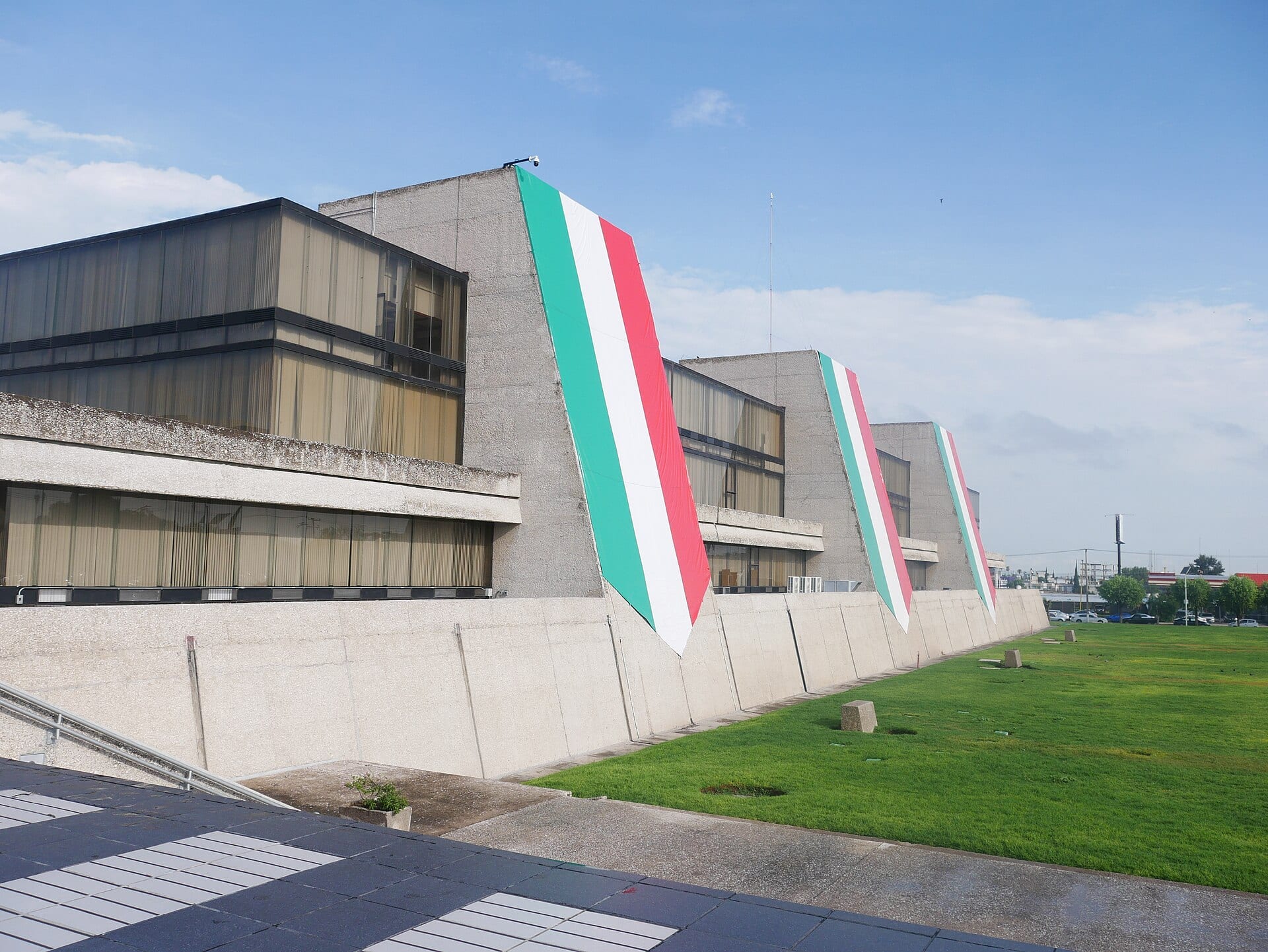 Fachada de concreto del edificio central del INEGI con tres banderas de México colgadas en sus muros inclinados, junto a un jardín.