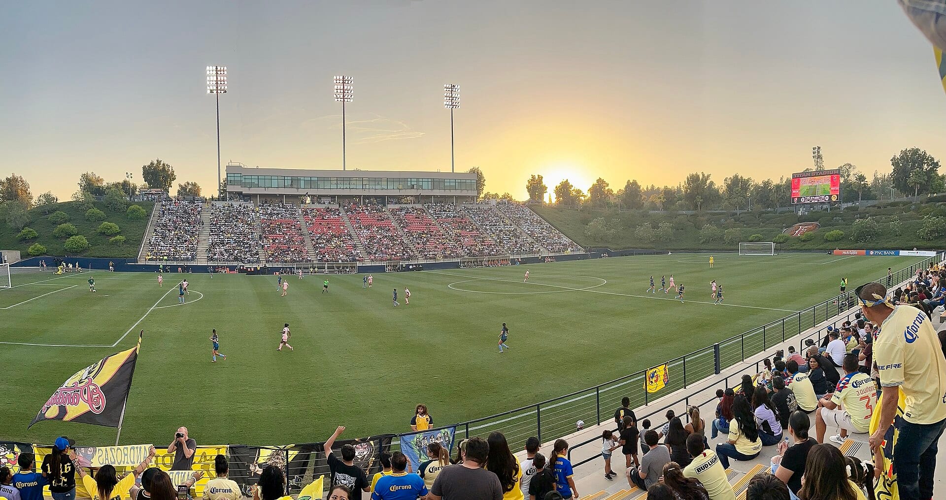 Panorámica de un partido de América Femenil al atardecer, con jugadoras en la cancha, tribunas llenas y aficionados con camisetas amarillas en primer plano.