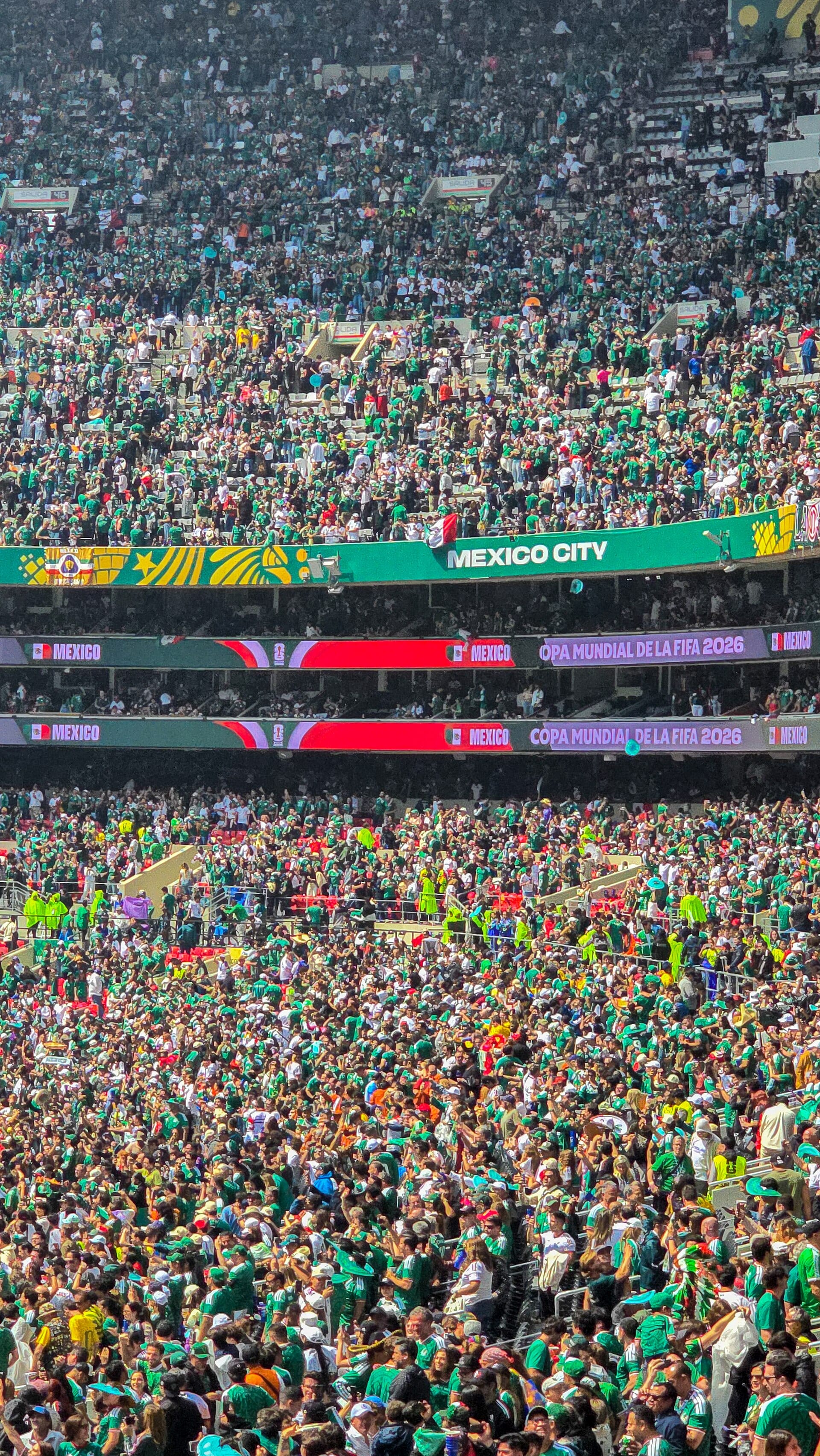 Gradas llenas de aficionados con camisetas verdes en el estadio Azteca, bajo un letrero que dice Mexico City.