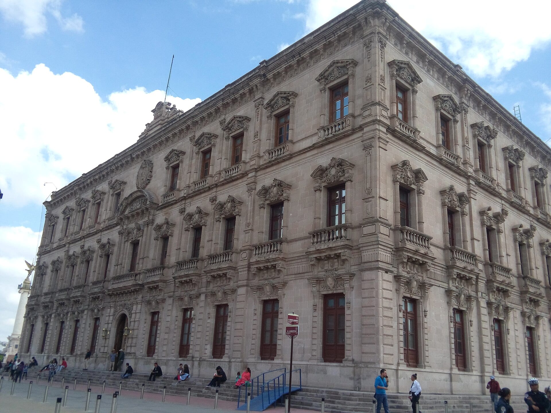 Esquina del Palacio de Gobierno de Chihuahua, edificio de cantera de tres pisos con balcones, y personas sentadas en la escalinata.