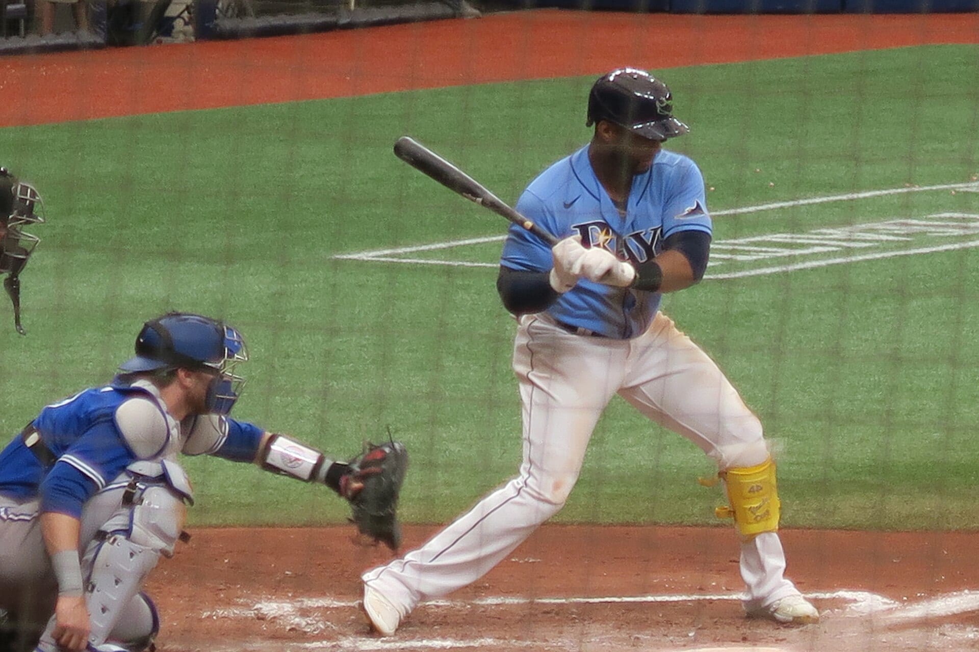 Yandy Díaz, con uniforme azul claro de los Rays, espera un lanzamiento en el plato junto al receptor rival, visto a través de una red.