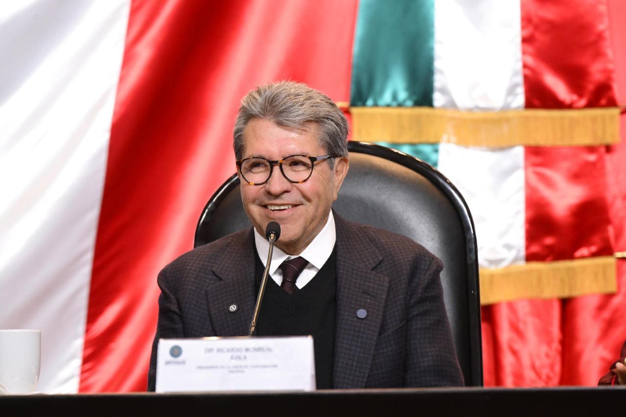 Ricardo Monreal sonríe sentado frente a un micrófono, con una placa con su nombre y la bandera de México al fondo.