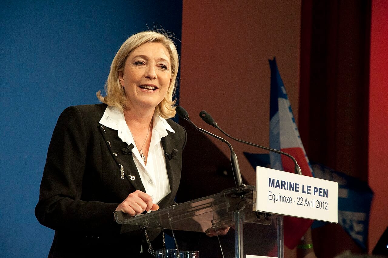 Marine Le Pen habla de pie ante un atril con micrófonos, con banderas de Francia detrás.