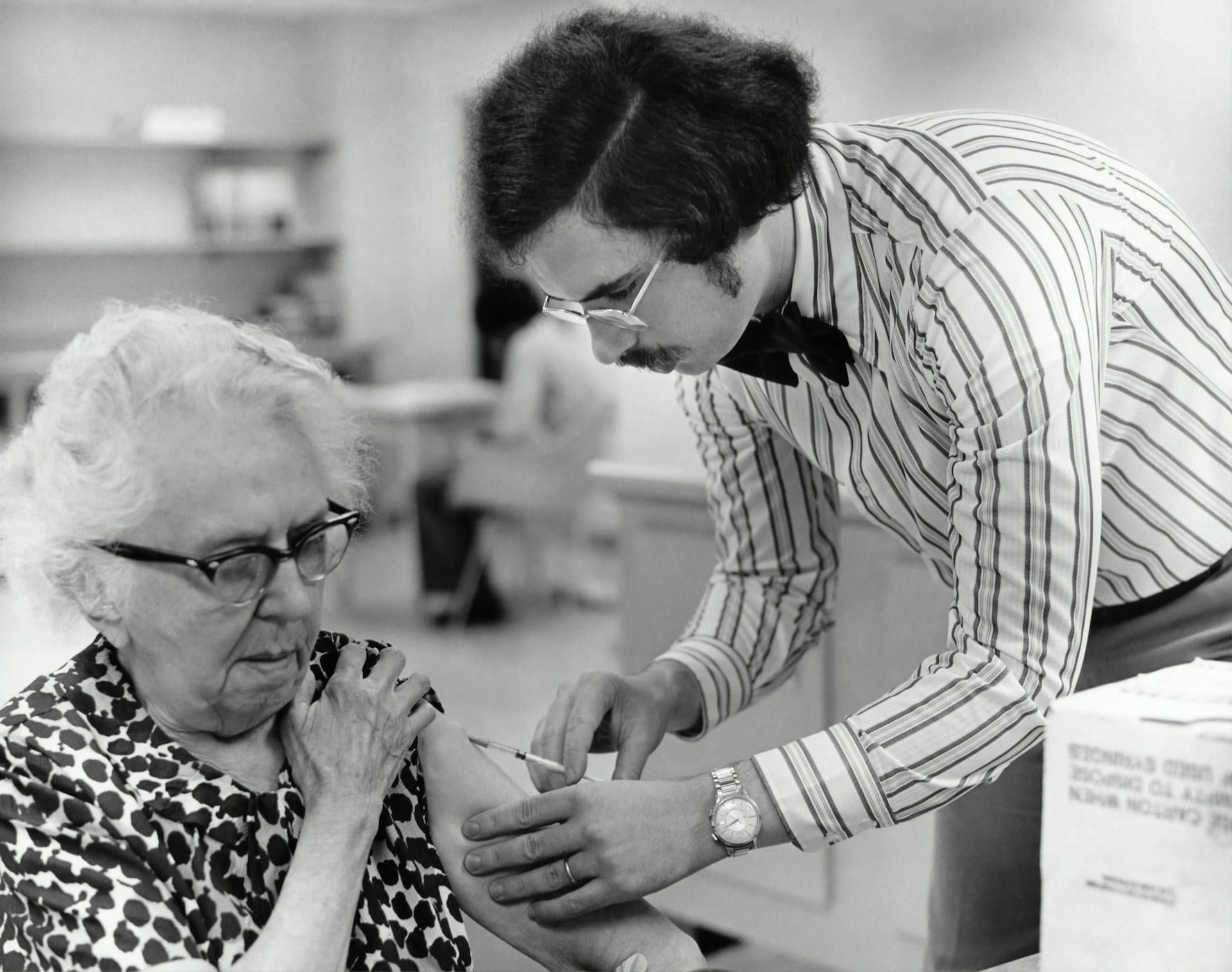 Fotografía en blanco y negro de un hombre con bigote que inyecta el brazo de una mujer mayor con gafas.