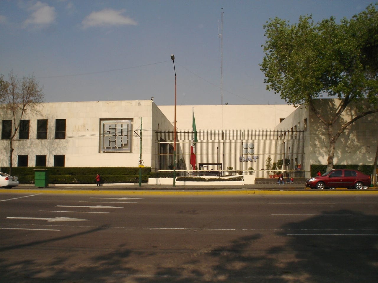 Fachada blanca de un edificio de oficinas con el logotipo de la Secretaría de Hacienda en el muro y las letras SAT tras una reja, vistas desde el otro lado de la avenida.
