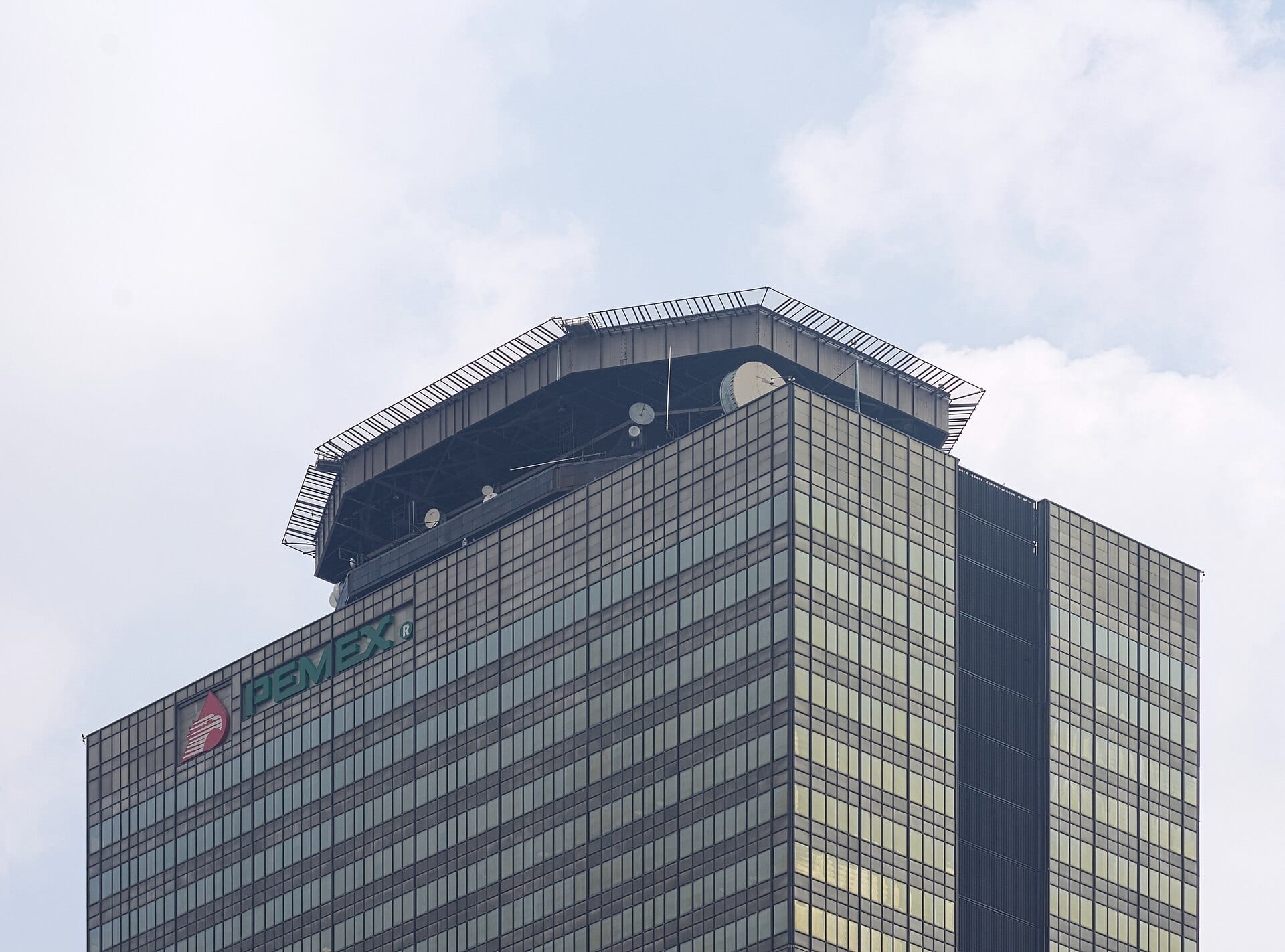 Parte alta de la Torre de Pemex en la Ciudad de México, un edificio de cristal con el logotipo de la empresa, visto desde abajo contra un cielo nublado.