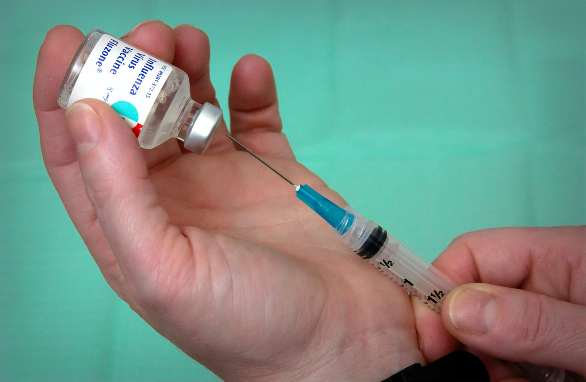 Una mano sostiene un vial de vacuna y una jeringa con aguja lista para aplicarla, sobre un fondo verde turquesa.