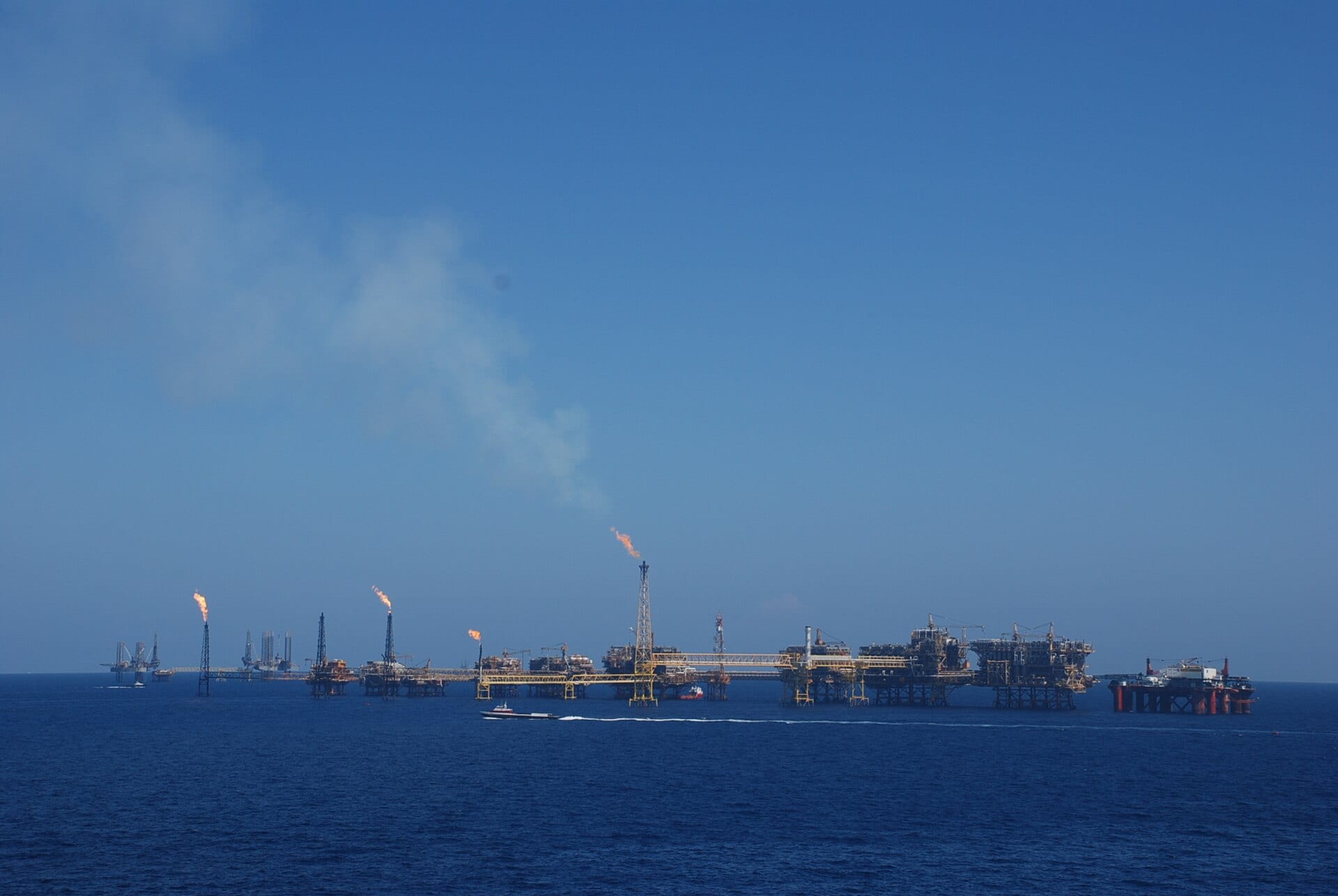 Plataformas petroleras unidas por pasarelas amarillas en mar abierto, con varias antorchas de gas encendidas y una lancha en primer plano.