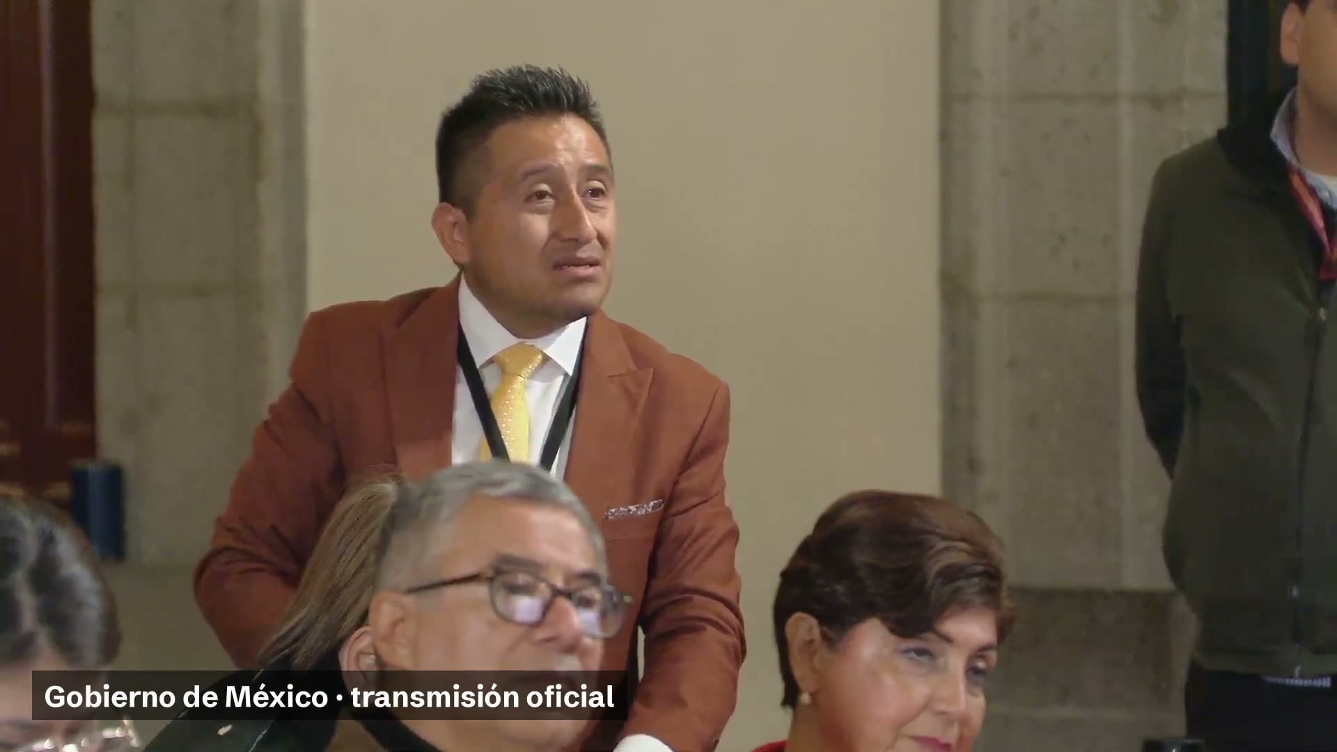 Un hombre de saco color teja y corbata amarilla, de pie entre los asistentes a la conferencia matutina.
