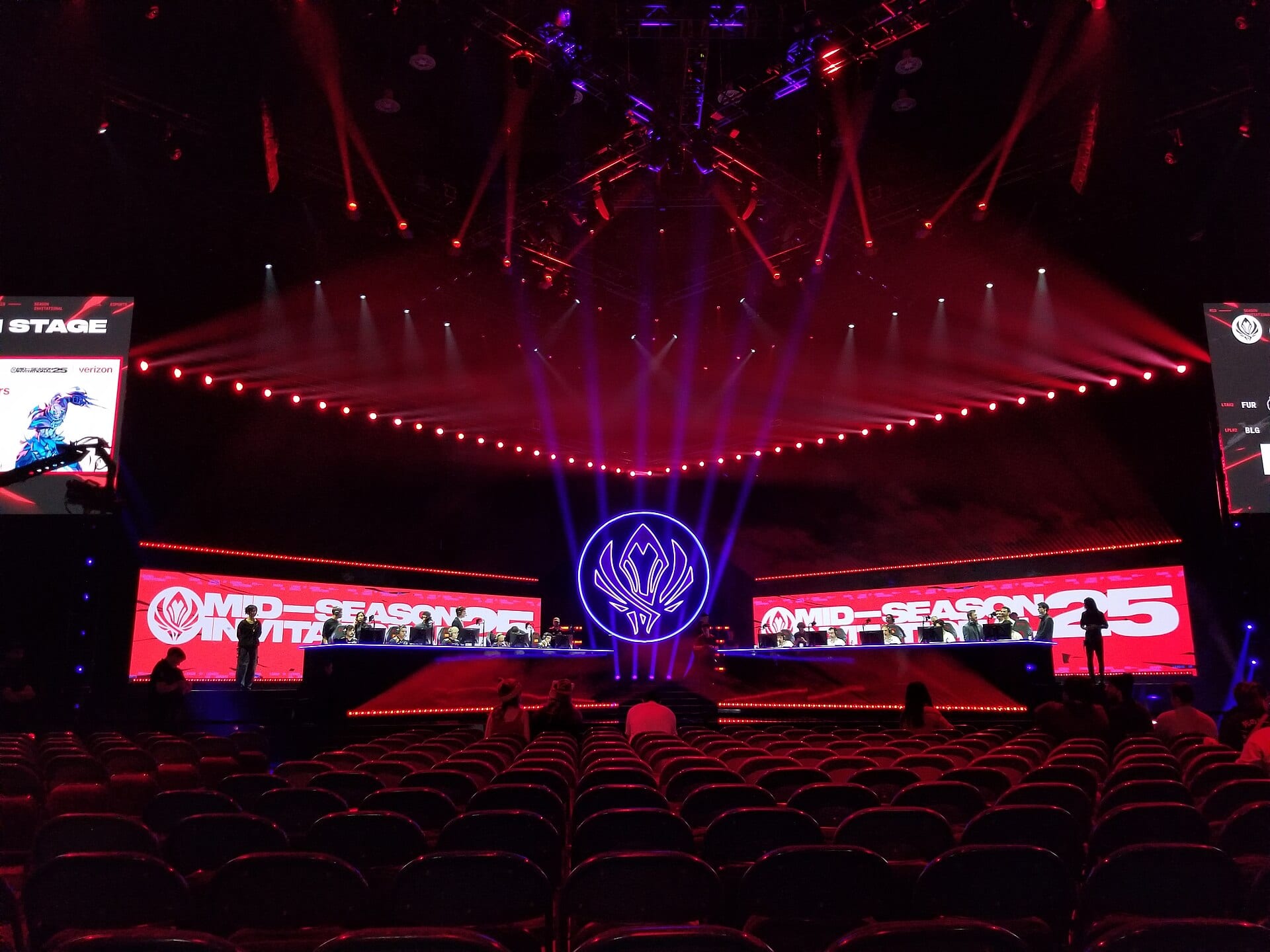 A League of Legends tournament stage with red screens and purple lights, players from two teams at their desks and a nearly empty arena in front.
