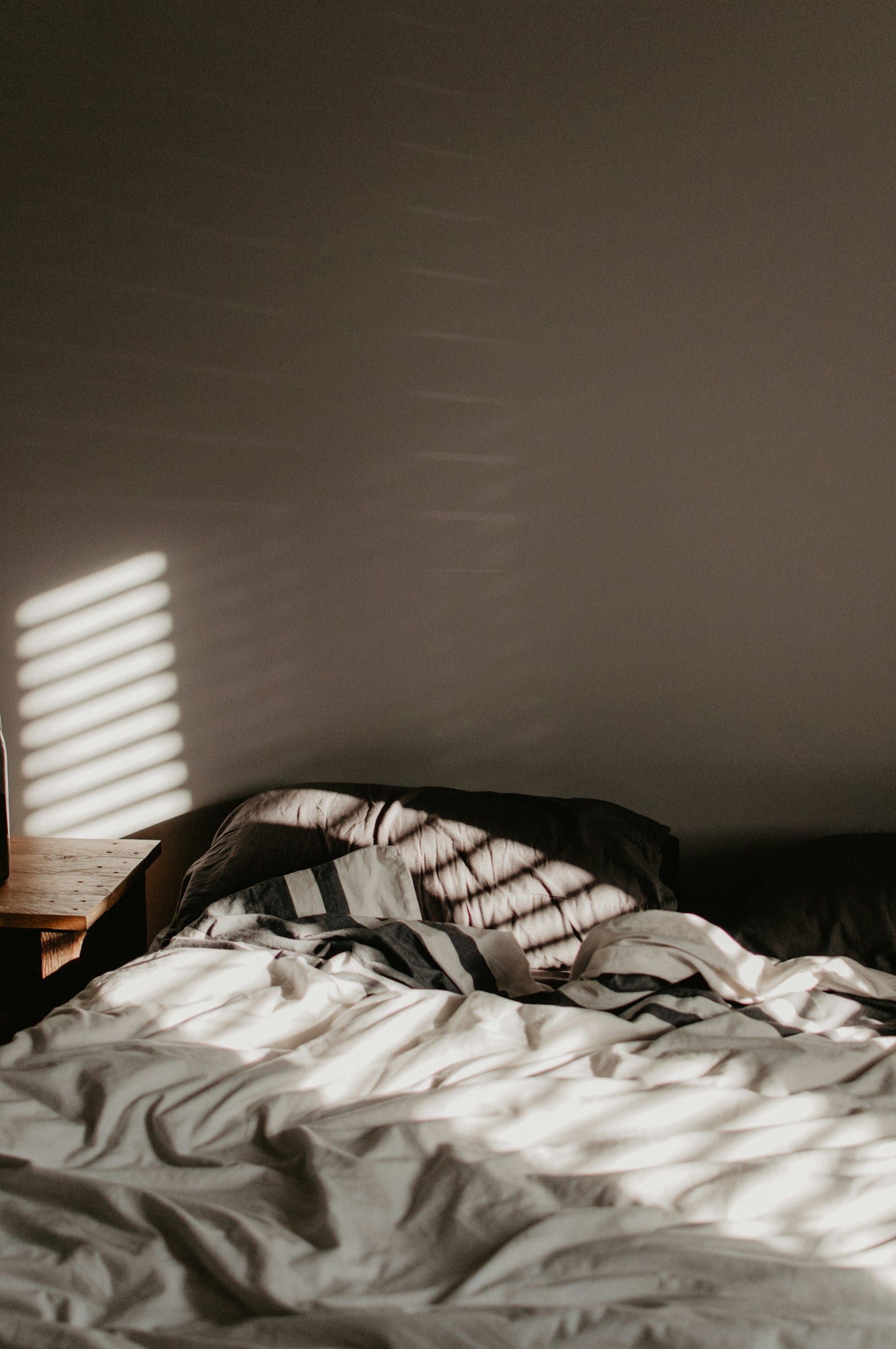 Una cama deshecha con sábanas blancas y rayas, con franjas de luz de una persiana sobre la pared y la almohada.
