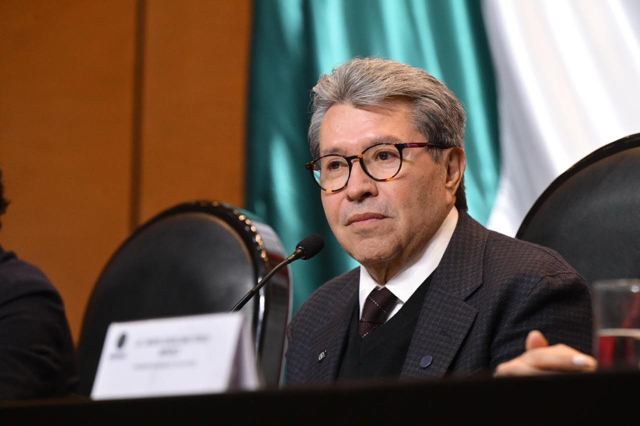 Ricardo Monreal, de lentes y traje oscuro, sentado frente a un micrófono con una bandera de México detrás.