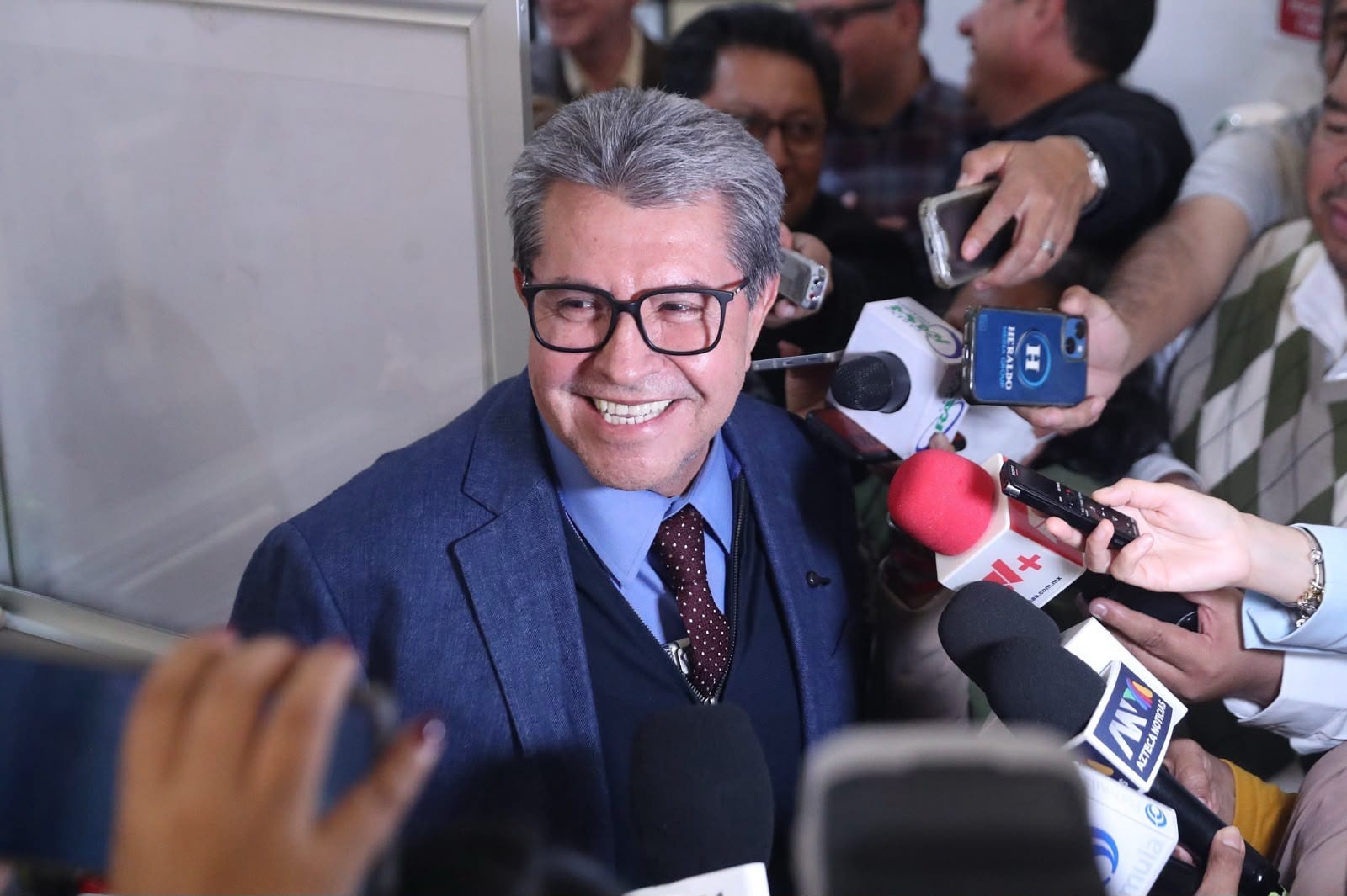 Ricardo Monreal sonríe con saco azul y corbata de lunares mientras reporteros le acercan micrófonos y grabadoras en un pasillo.
