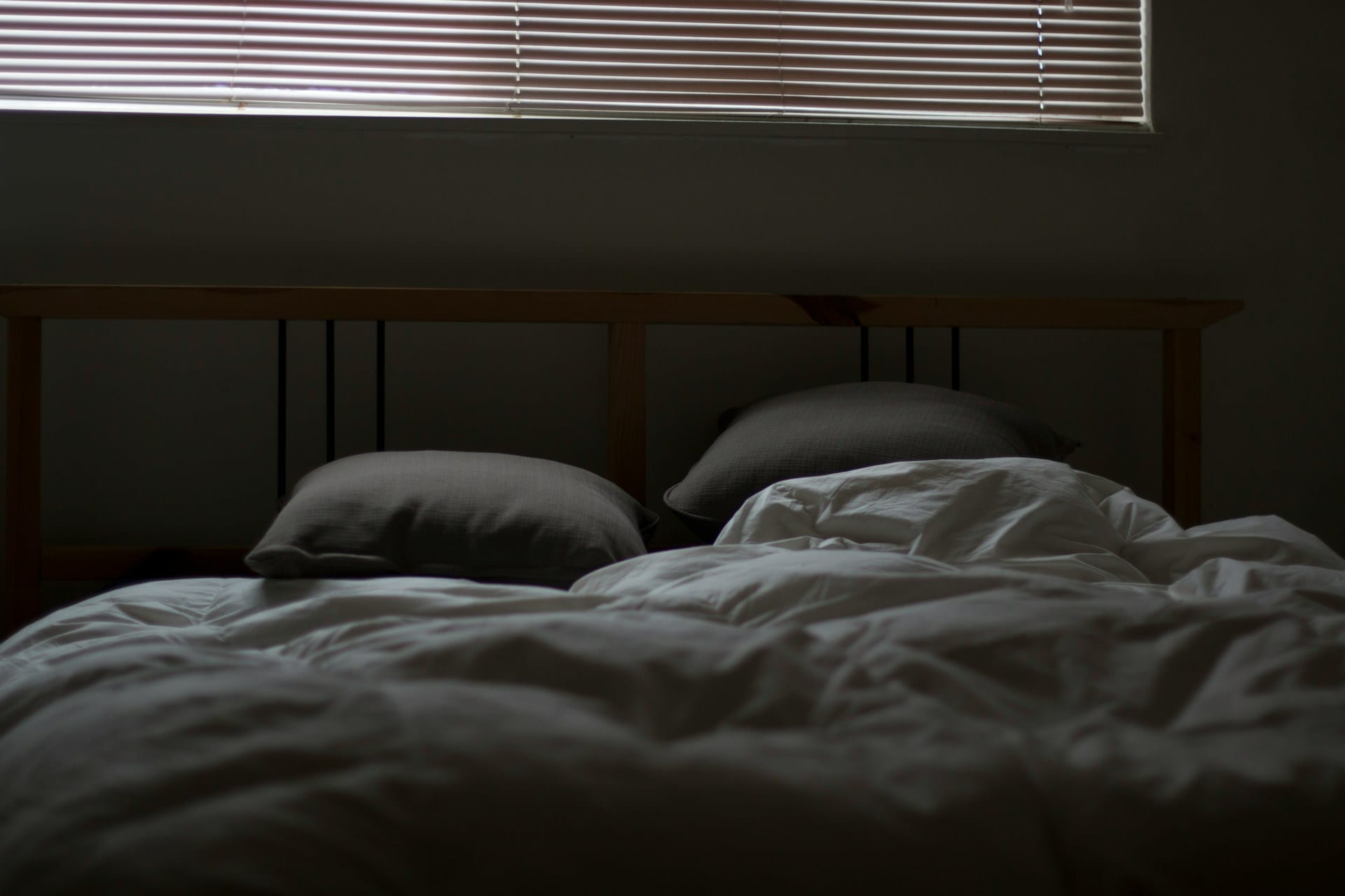 Una cama deshecha con edredón blanco y dos almohadas grises en un cuarto en penumbra, con la luz de una persiana al fondo.