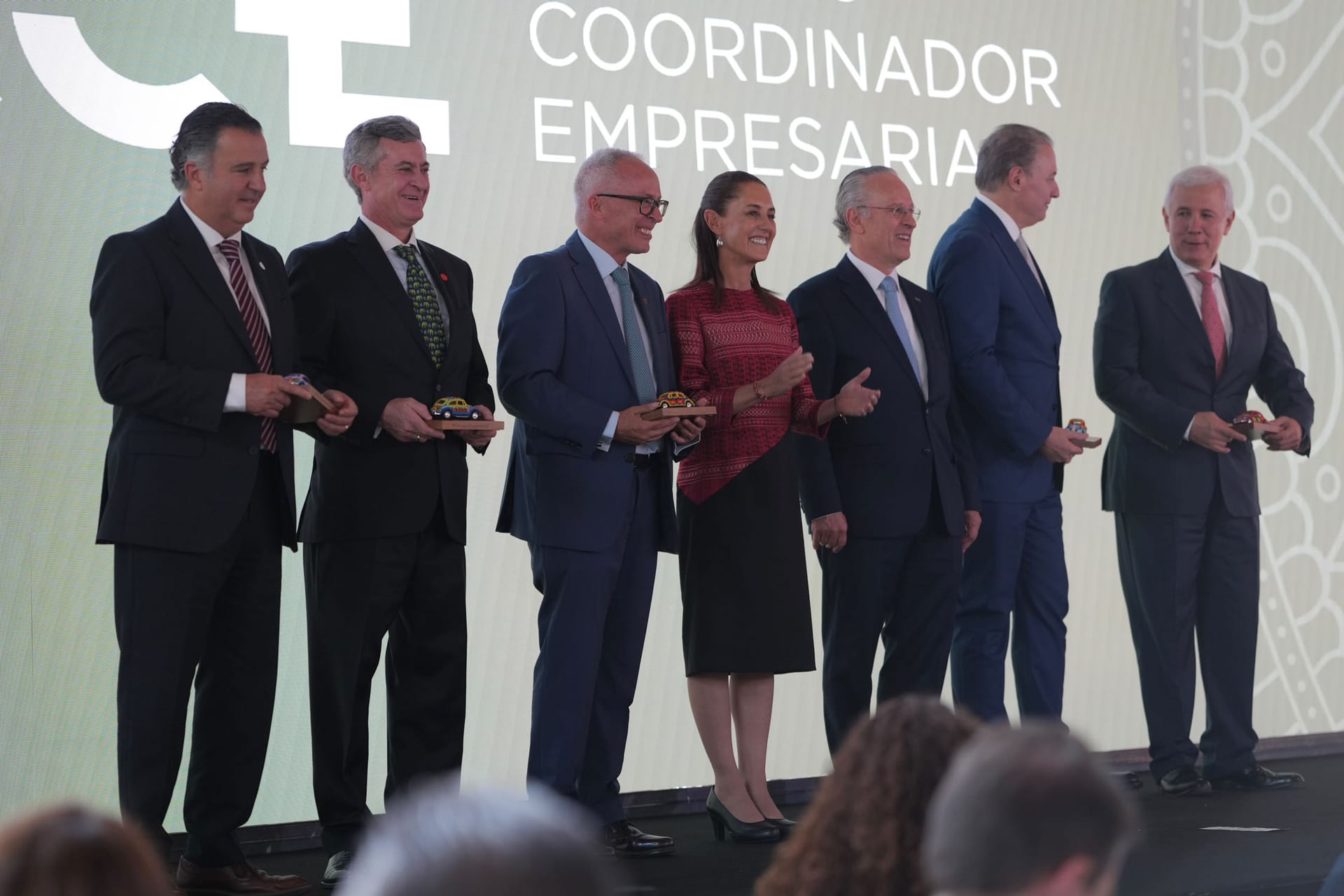 Claudia Sheinbaum sonríe y aplaude en un escenario junto a seis empresarios que sostienen autos de juguete de colores, frente a una pantalla del CCE.