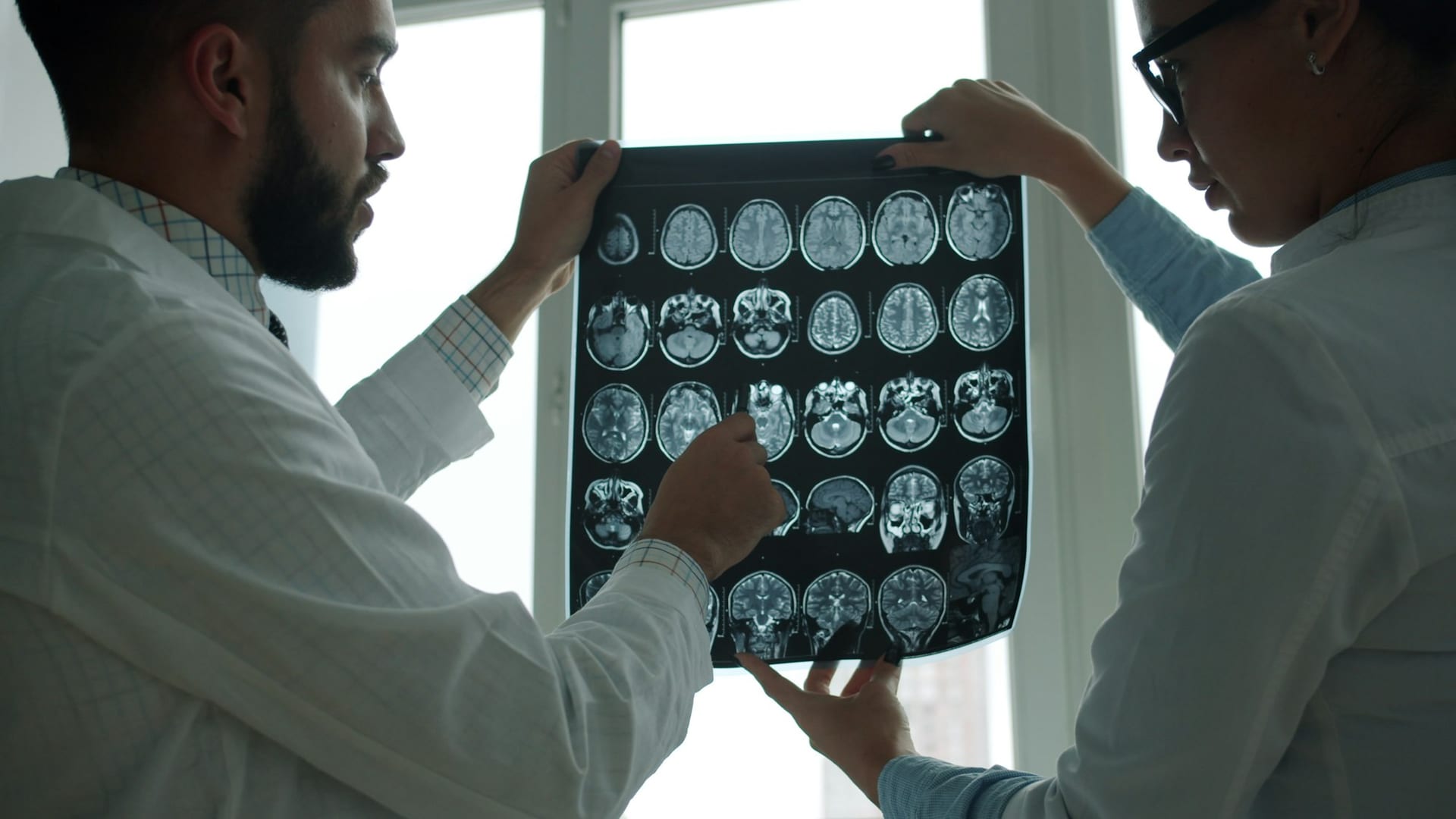 Una médica y un médico sostienen a contraluz una placa con varias imágenes de resonancia magnética del cerebro.