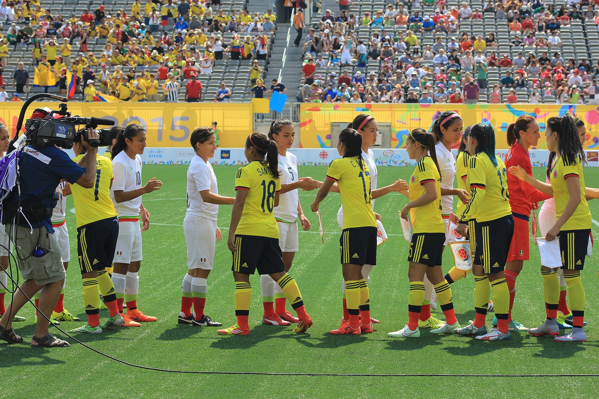 Jugadoras de México, de blanco, y de Colombia, de amarillo, se saludan en fila sobre el césped antes de un partido, con un camarógrafo a la izquierda.