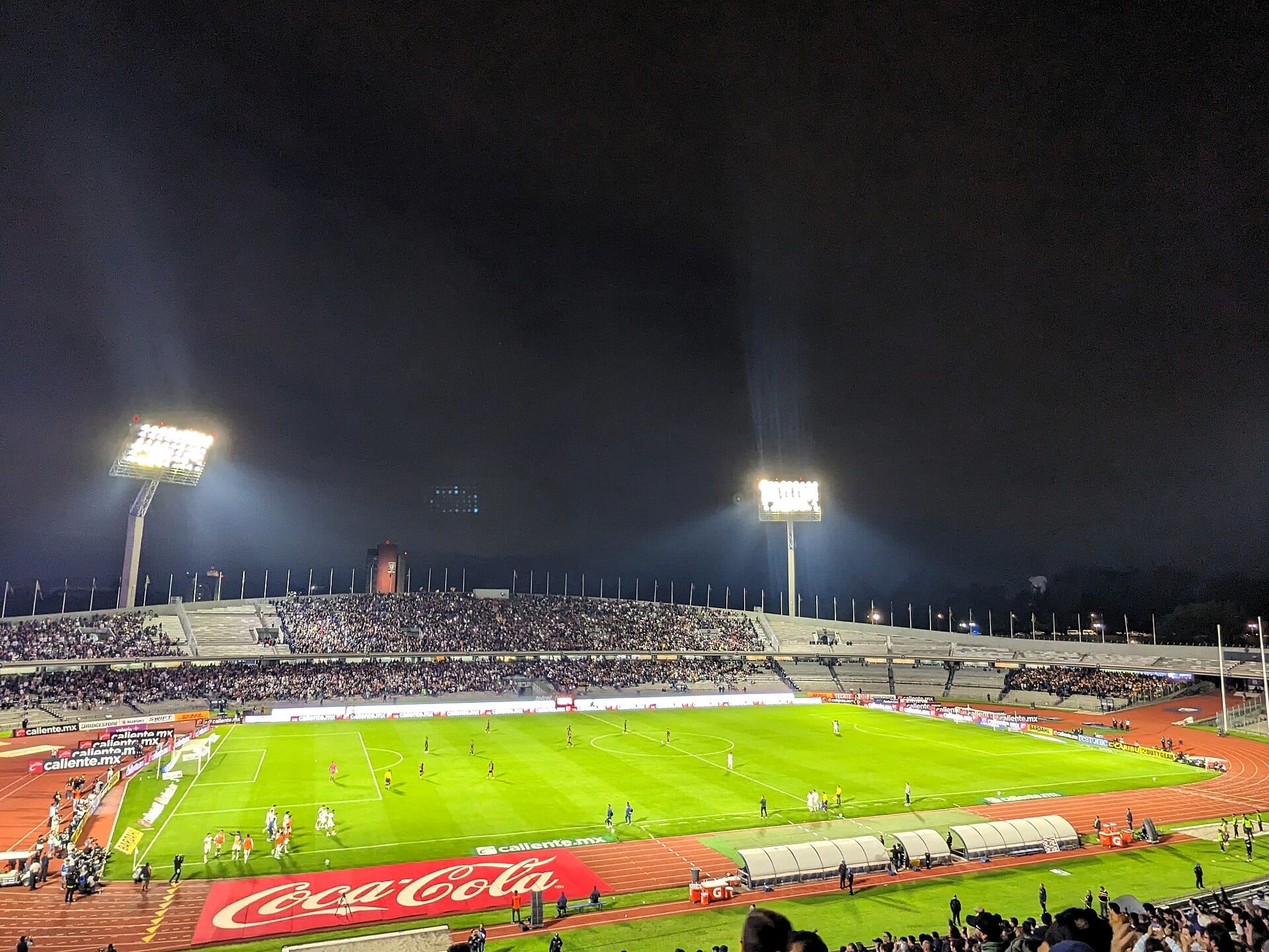Vista nocturna del Estadio Olímpico Universitario con las gradas llenas, dos torres de luz encendidas y jugadores dispersos sobre la cancha.
