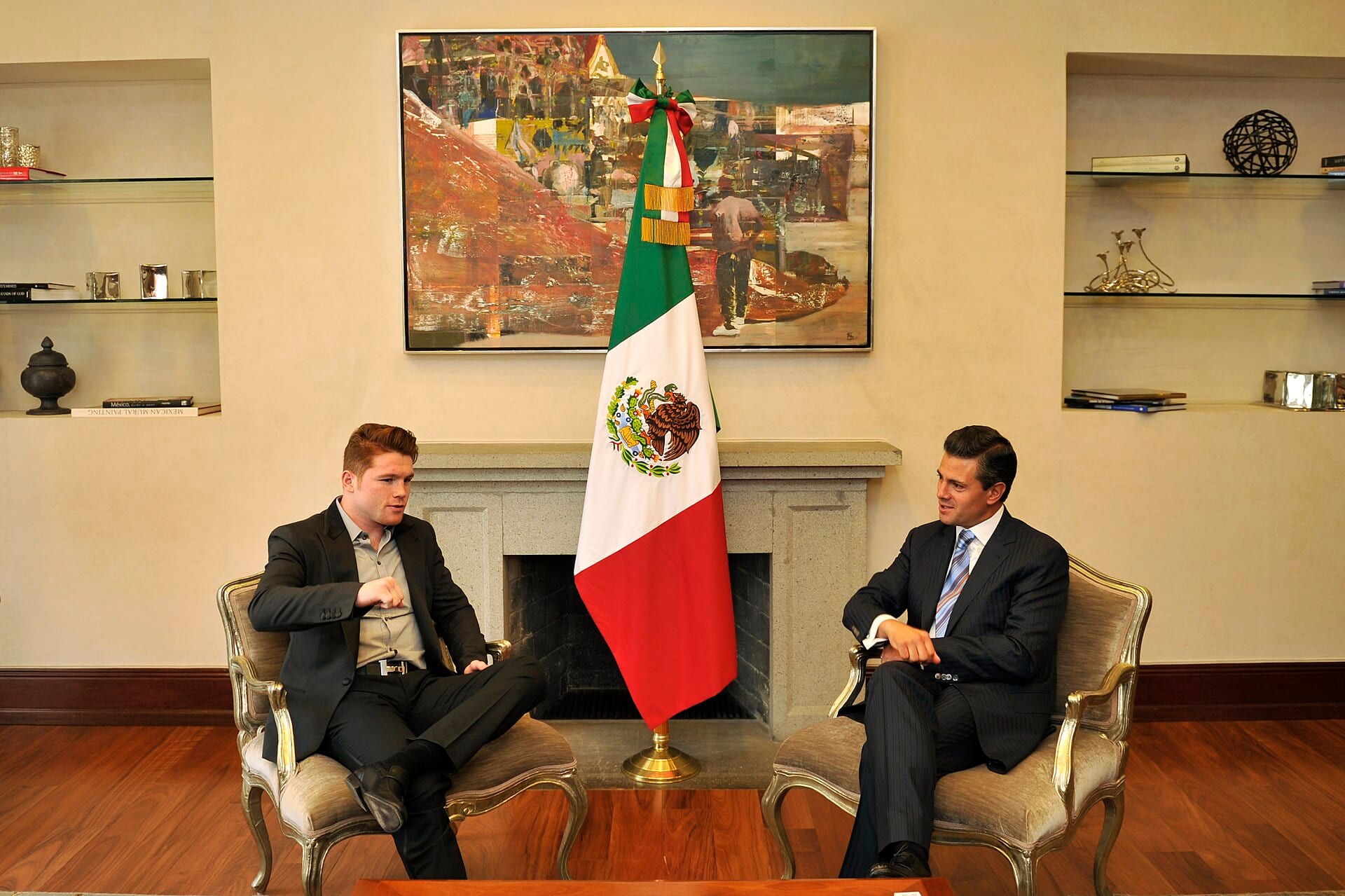 Saúl Álvarez, de saco oscuro, conversa sentado en un sillón junto a otro hombre de traje, con una bandera de México entre ambos.