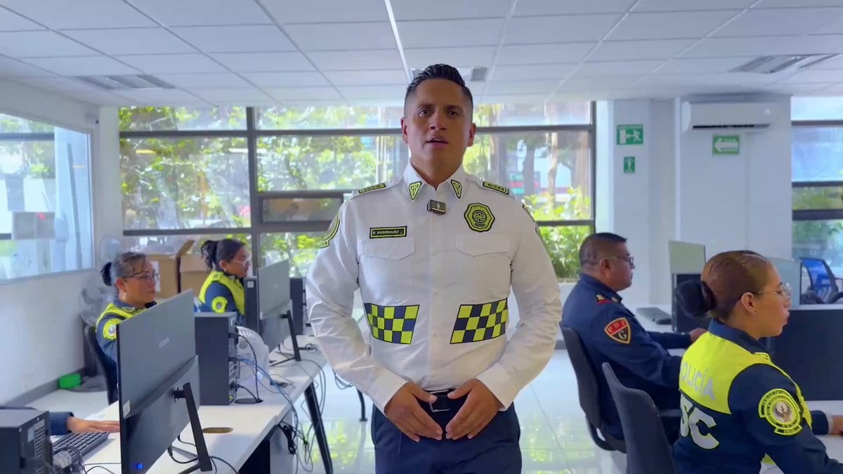 Un oficial de la Subsecretaría de Control de Tránsito de la SSC, de camisa blanca con cuadros amarillos y negros, habla a cámara en una oficina donde otros policías trabajan frente a sus computadoras.