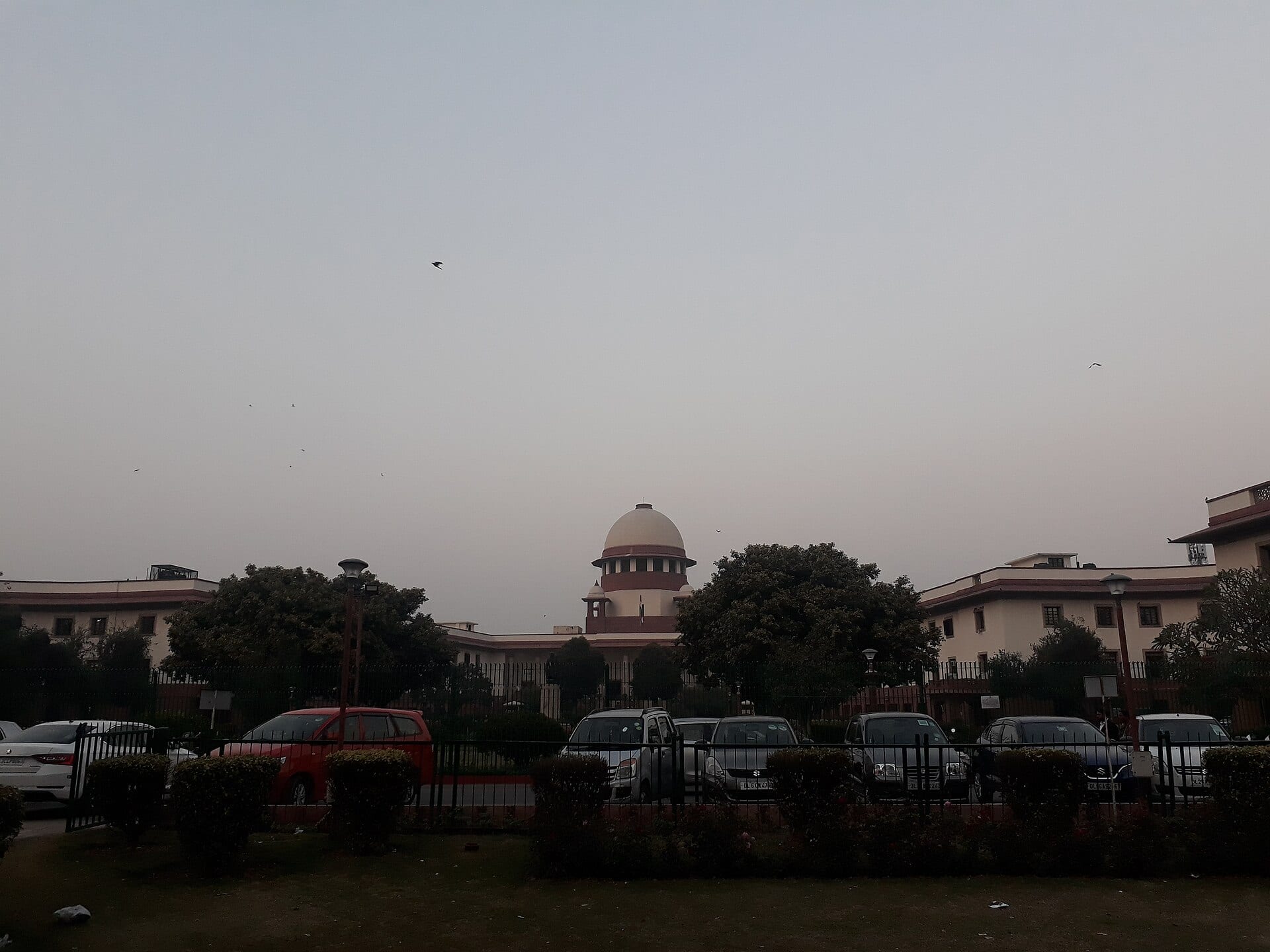 La cúpula blanca y roja del edificio de la Corte Suprema de la India asoma tras los árboles y una reja, con autos estacionados al frente.