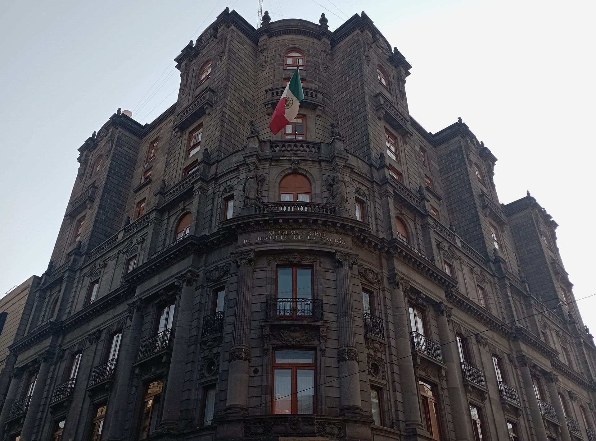 Esquina de un edificio de cantera oscura con balcones, una bandera de México y el letrero Suprema Corte de Justicia de la Nación, visto desde la calle.
